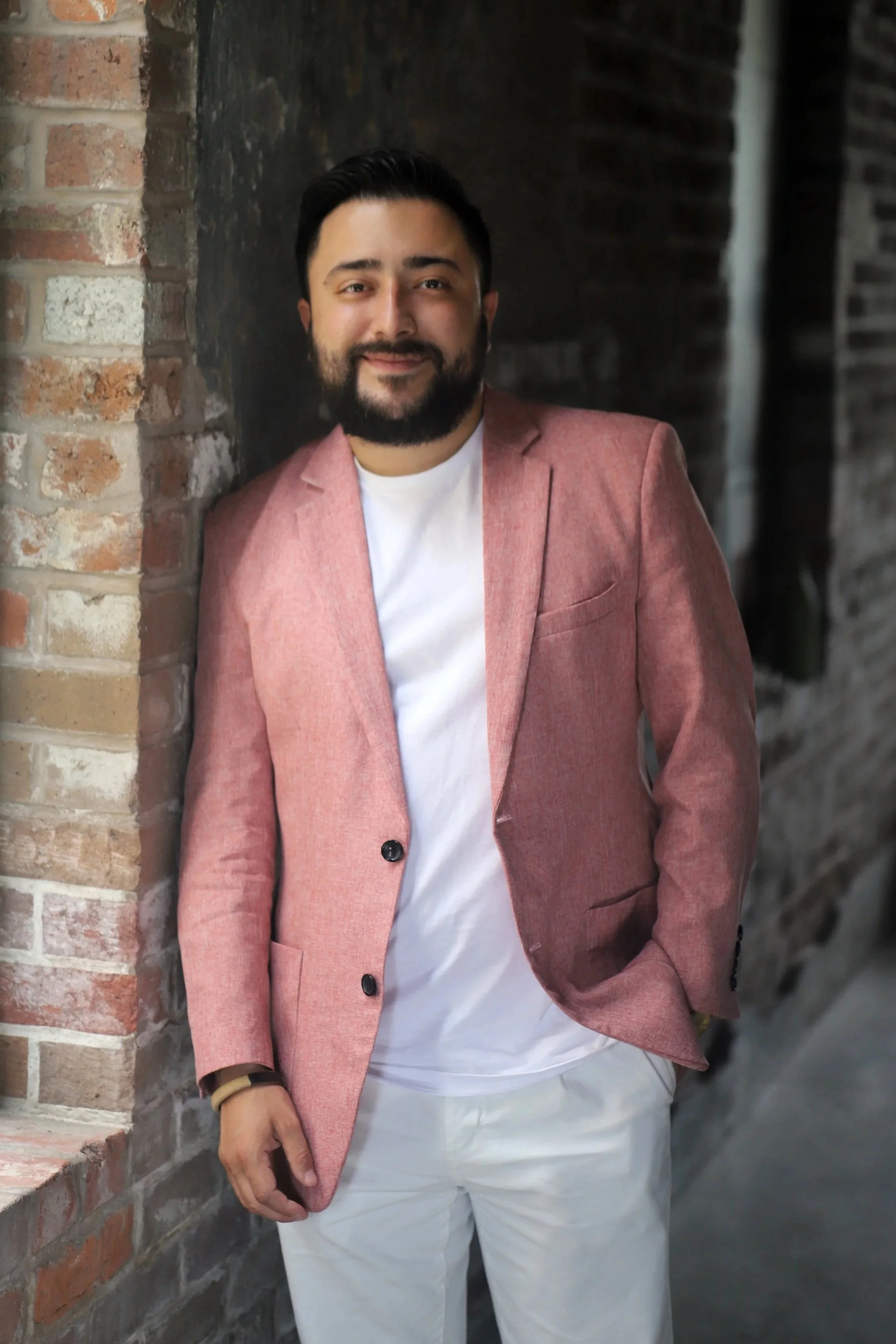 A man with dark hair, a beard, and earrings sitting in a modern black and wood chair in front of a brick wall. He is wearing a pink blazer, white shirt, white pants, and a gold wristwatch.