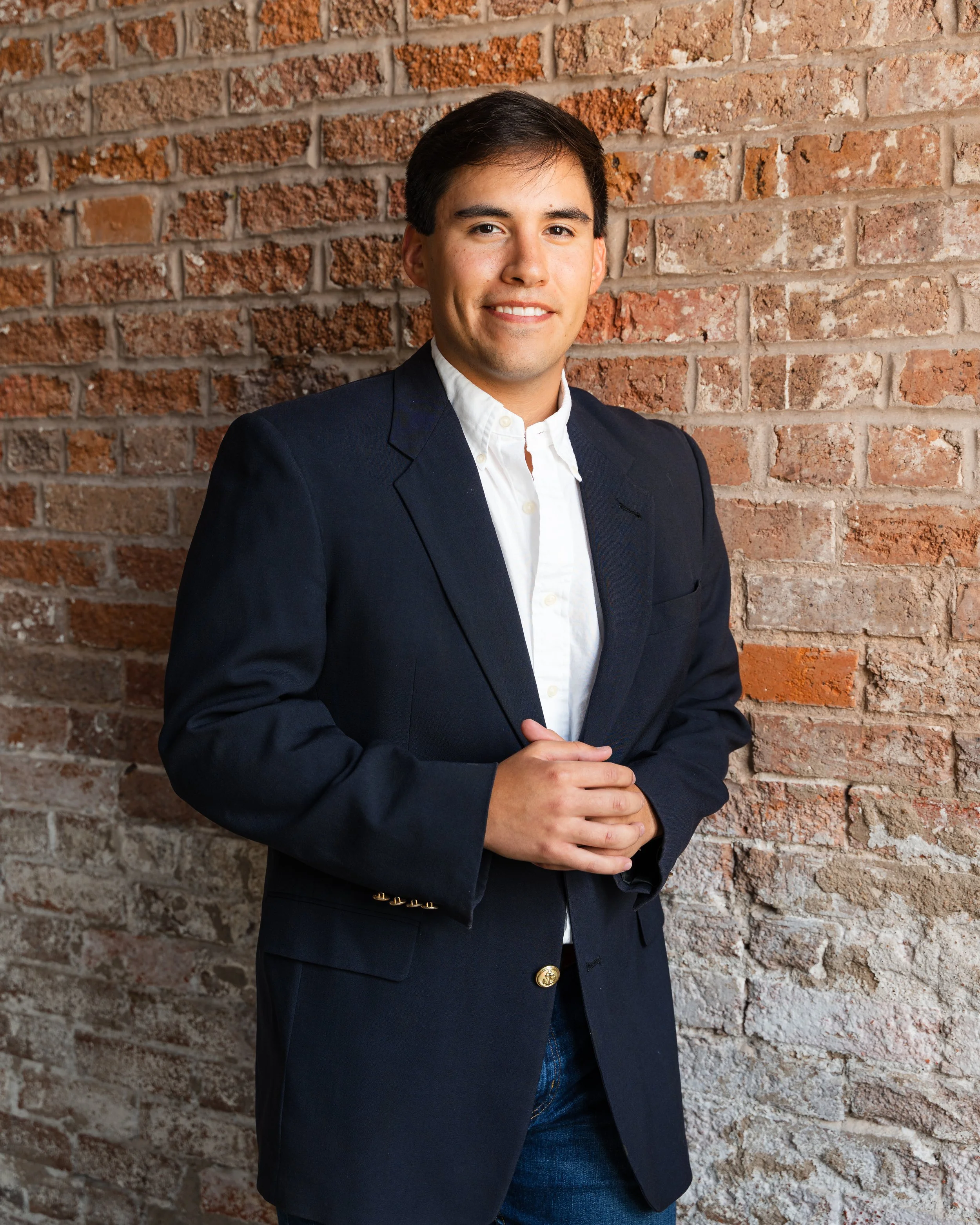 A young man with glasses and dark hair, dressed in a black button-up shirt with a small red logo, gray pants, and a brown belt, standing outdoors against a brick wall and smiling at the camera.