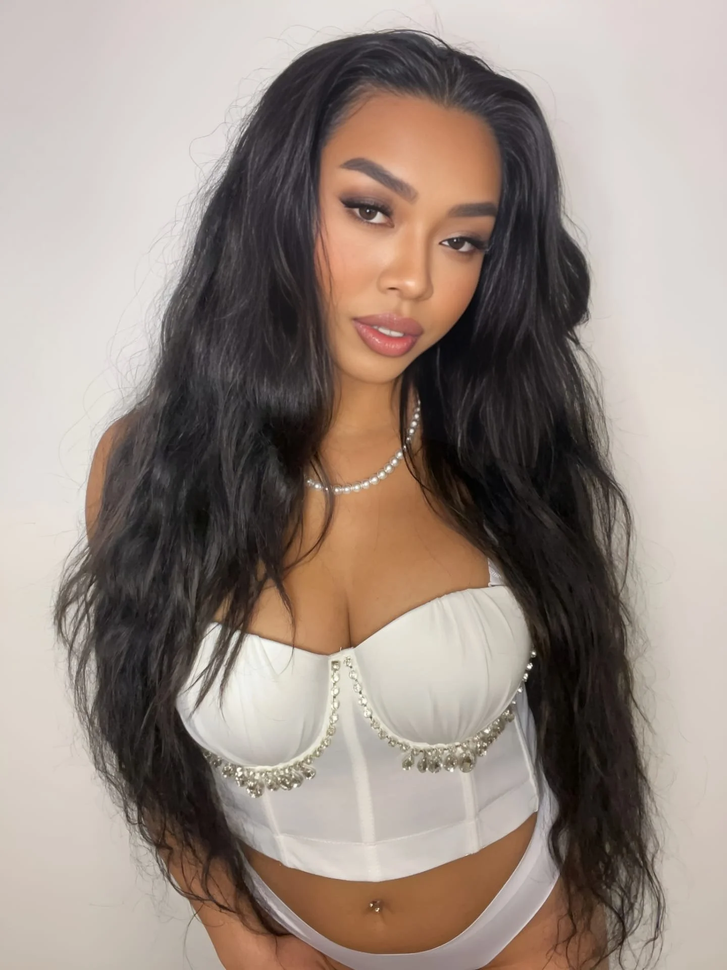 A woman with long, wavy black hair, wearing a white strapless top adorned with pearls and rhinestones, and a pearl necklace. She has makeup with defined eyebrows, eyeliner, and glossy lips, standing against a plain light background.