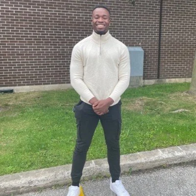 A smiling young man standing outdoors on a sidewalk with grass and a brick building in the background. He is wearing a light-colored zip-up sweater, dark pants, and white sneakers.