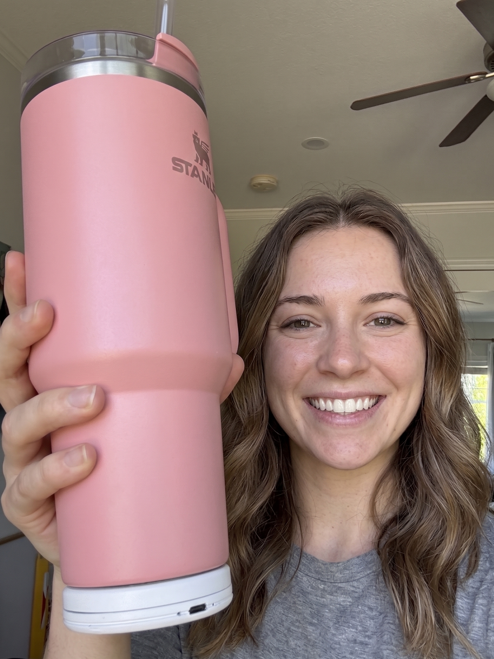 A smiling woman holding a large pink tumbler with a lid and straw which has its water intake tracked by the Sponge device, which is attached to the bottom. , standing inside a home with a ceiling fan and window in the background.