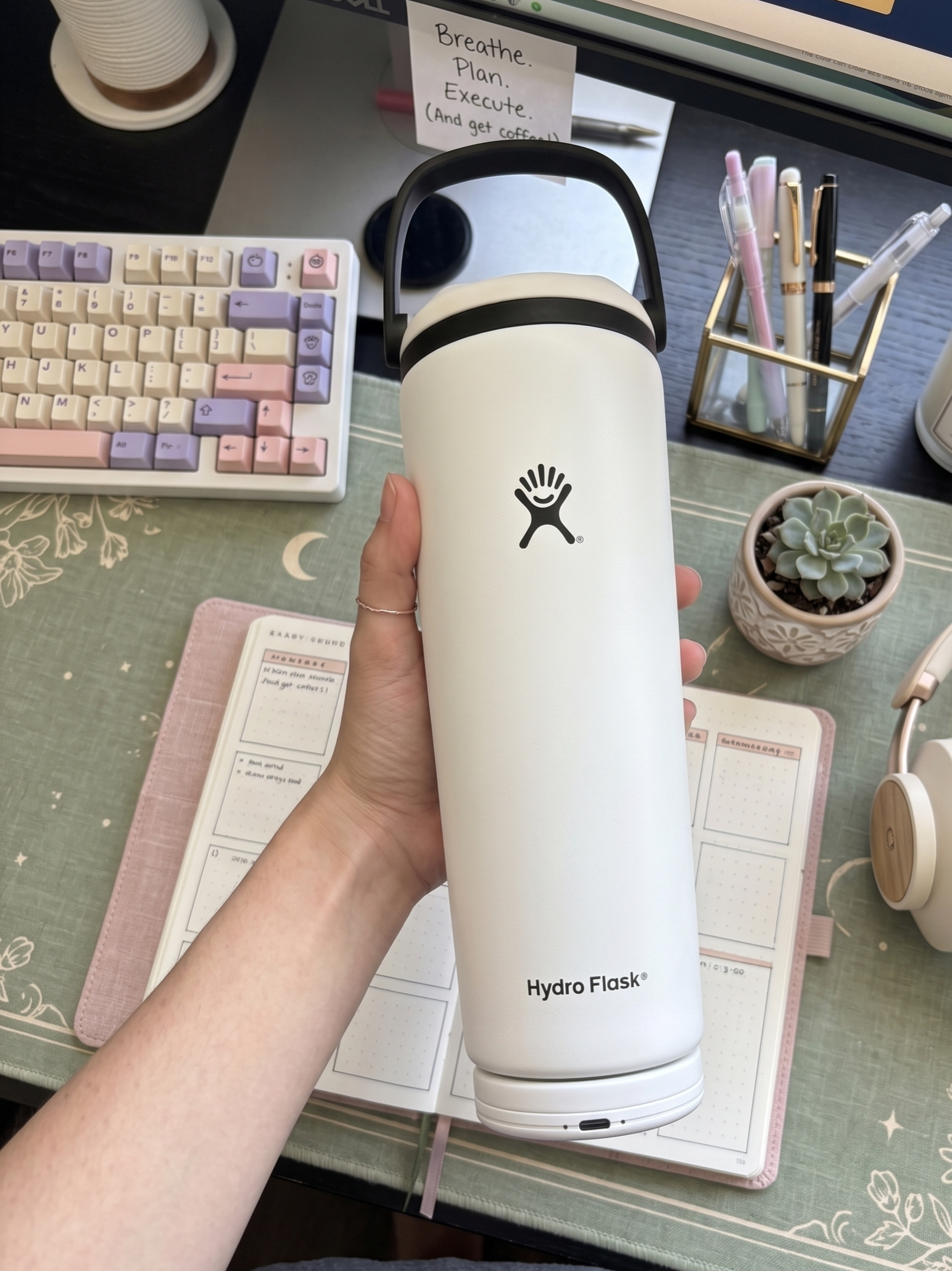 Close-up of a person's hand holding a white Hydro Flask water bottle with a Sponge Device snapped on the bottom. On the desk is a small potted succulent, a cup of coffee, and a note that says, "Breathe. Plan. Execute. (And get coffee)."