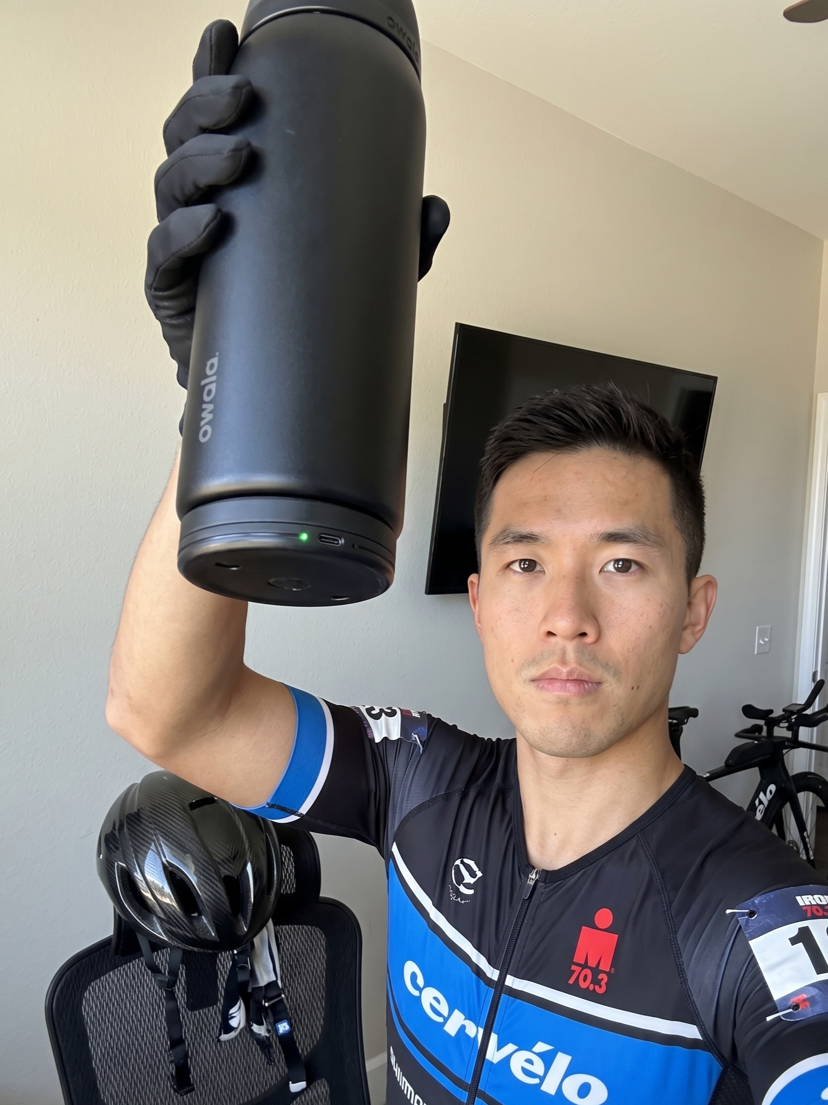 A young man in cycling gear holding a black insulated water bottle with his Sponge Device clipped to the bottom, standing indoors with a bike helmet hanging on a chair and a bicycle in the background.