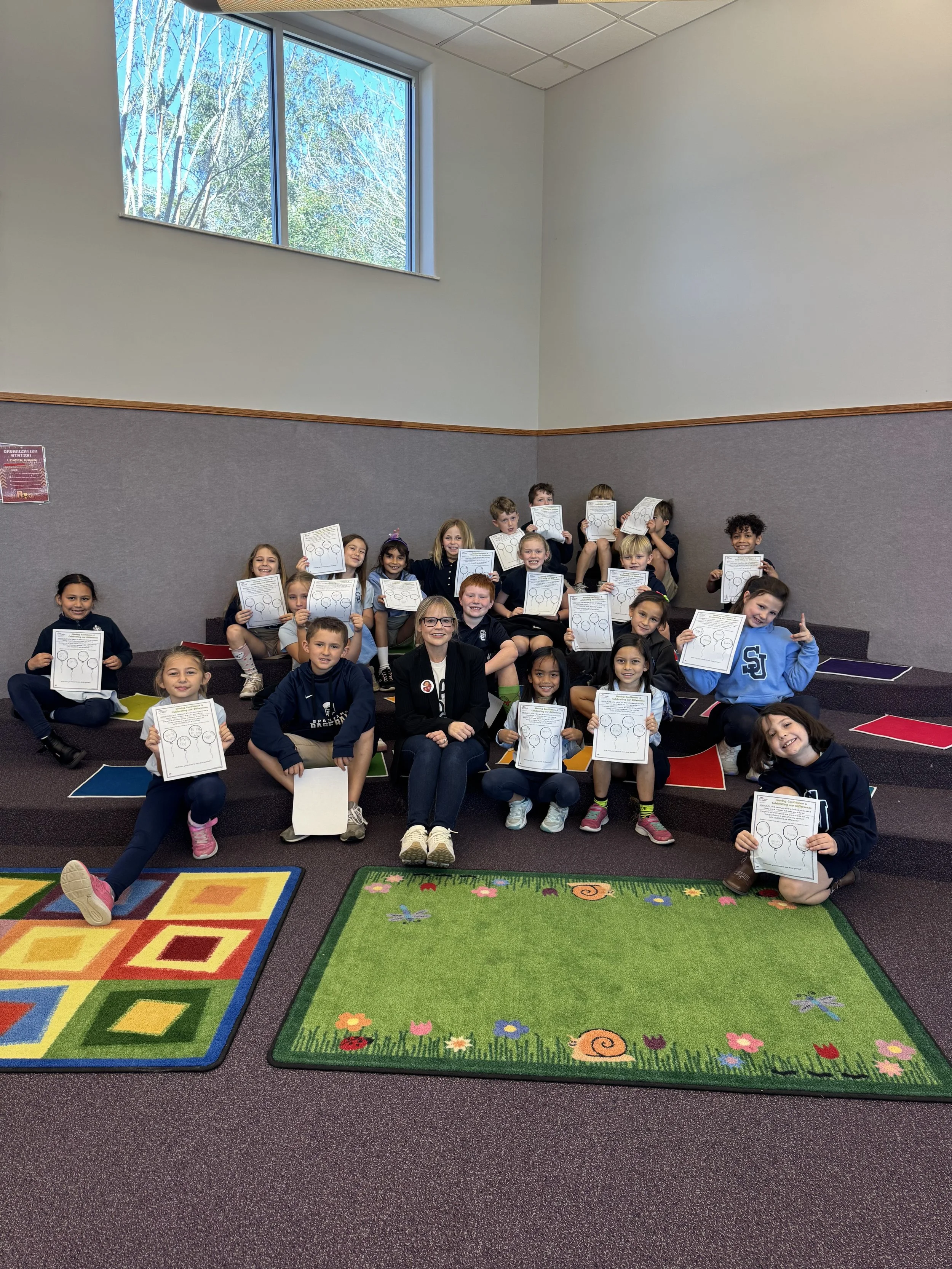 A group of schoolchildren and a teacher sitting on steps in a classroom, holding certificates and smiling. The room has a colorful carpet and a large window showing trees outside.