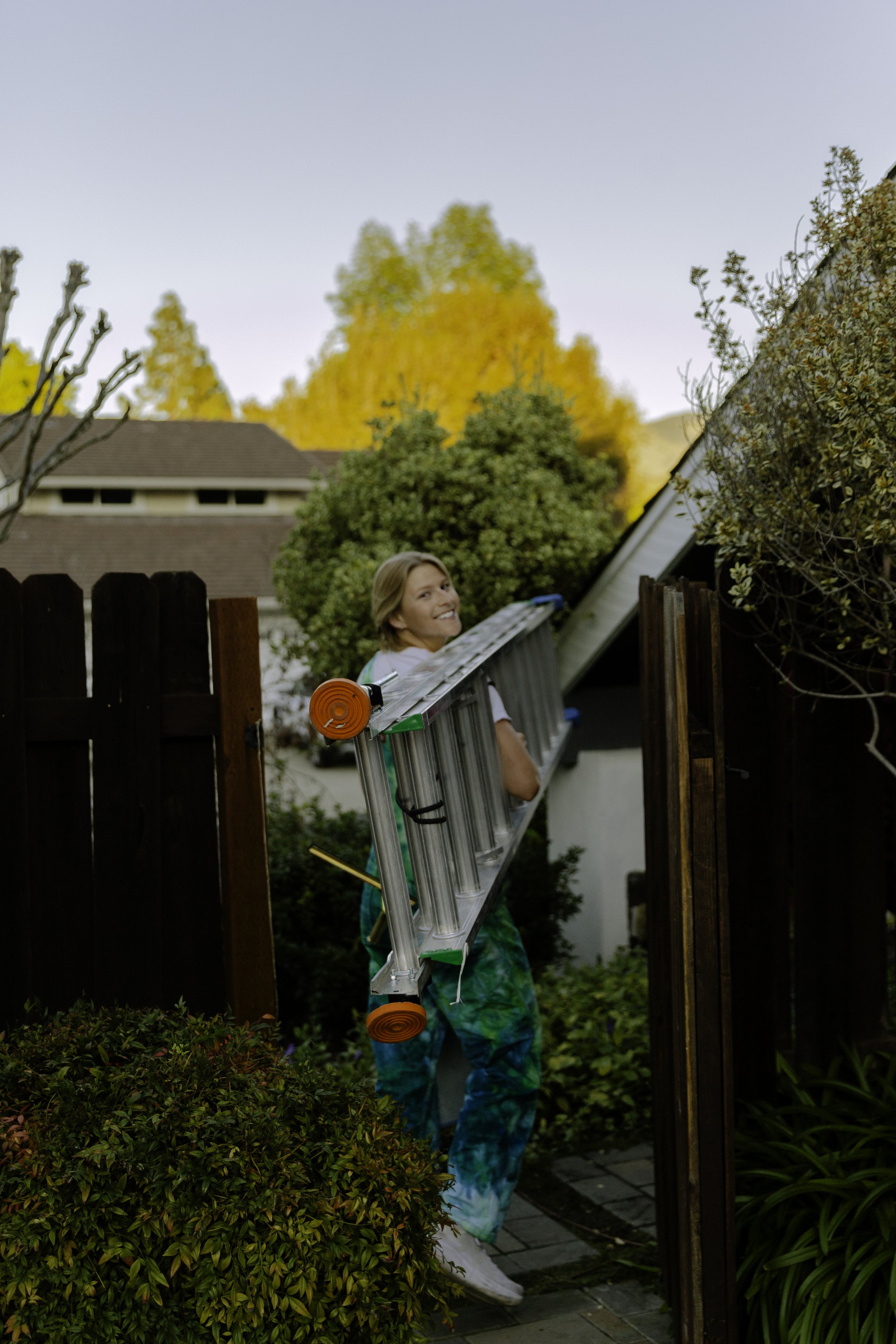 A woman carrying a ladder through a backyard gate with trees in autumn foliage in the background.
