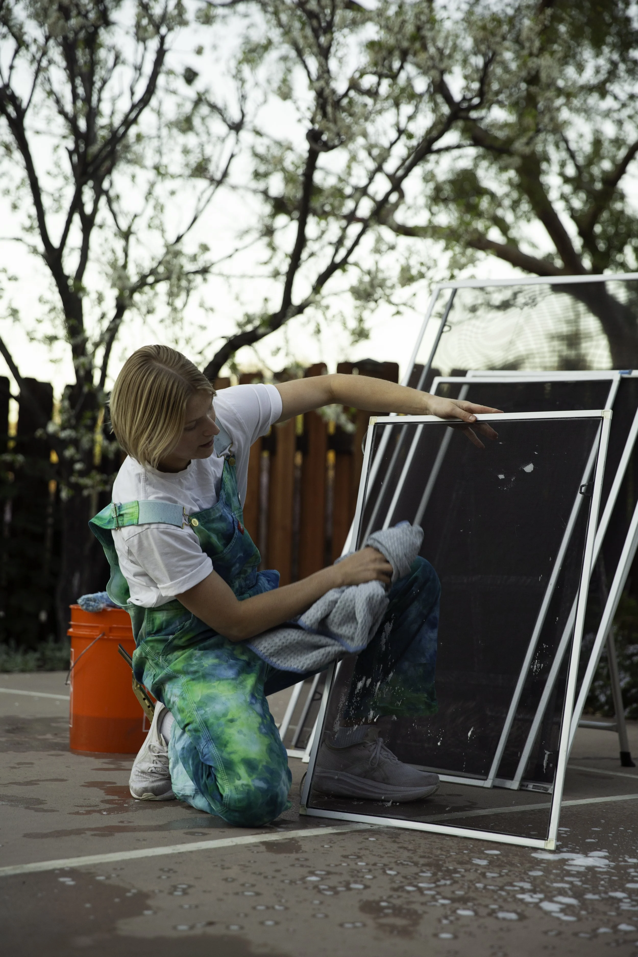 A young person cleaning multiple large glass or mirror panes outdoors during daytime. They are kneeling on the ground, wearing a white t-shirt and colorful tie-dye pants, with a bucket nearby and trees in the background.