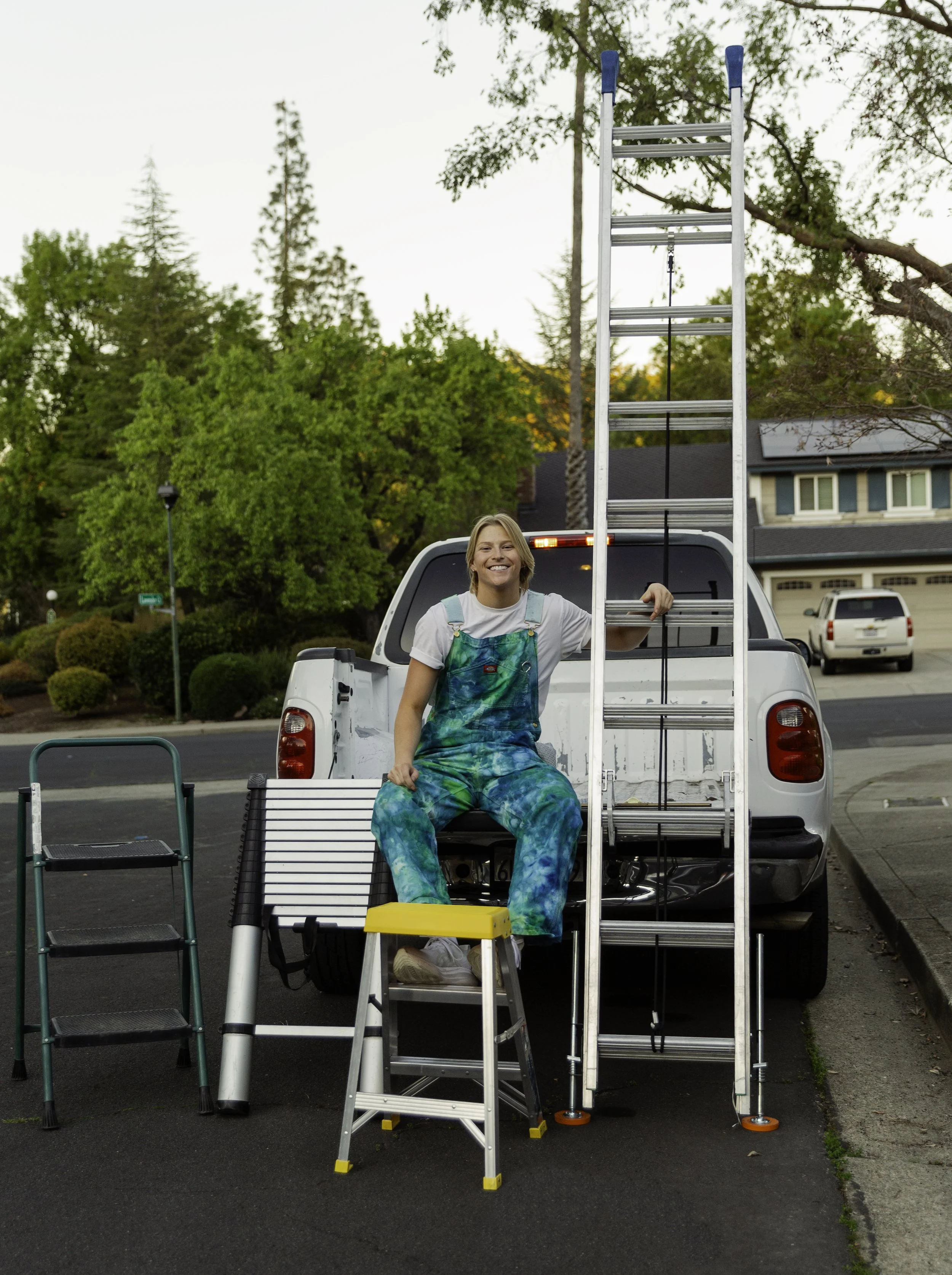A woman in colorful overalls sitting on the tailgate of a white pickup truck, smiling, with a ladder, step stool, and other ladder parts in front of her, outdoors in a suburban neighborhood during daytime.