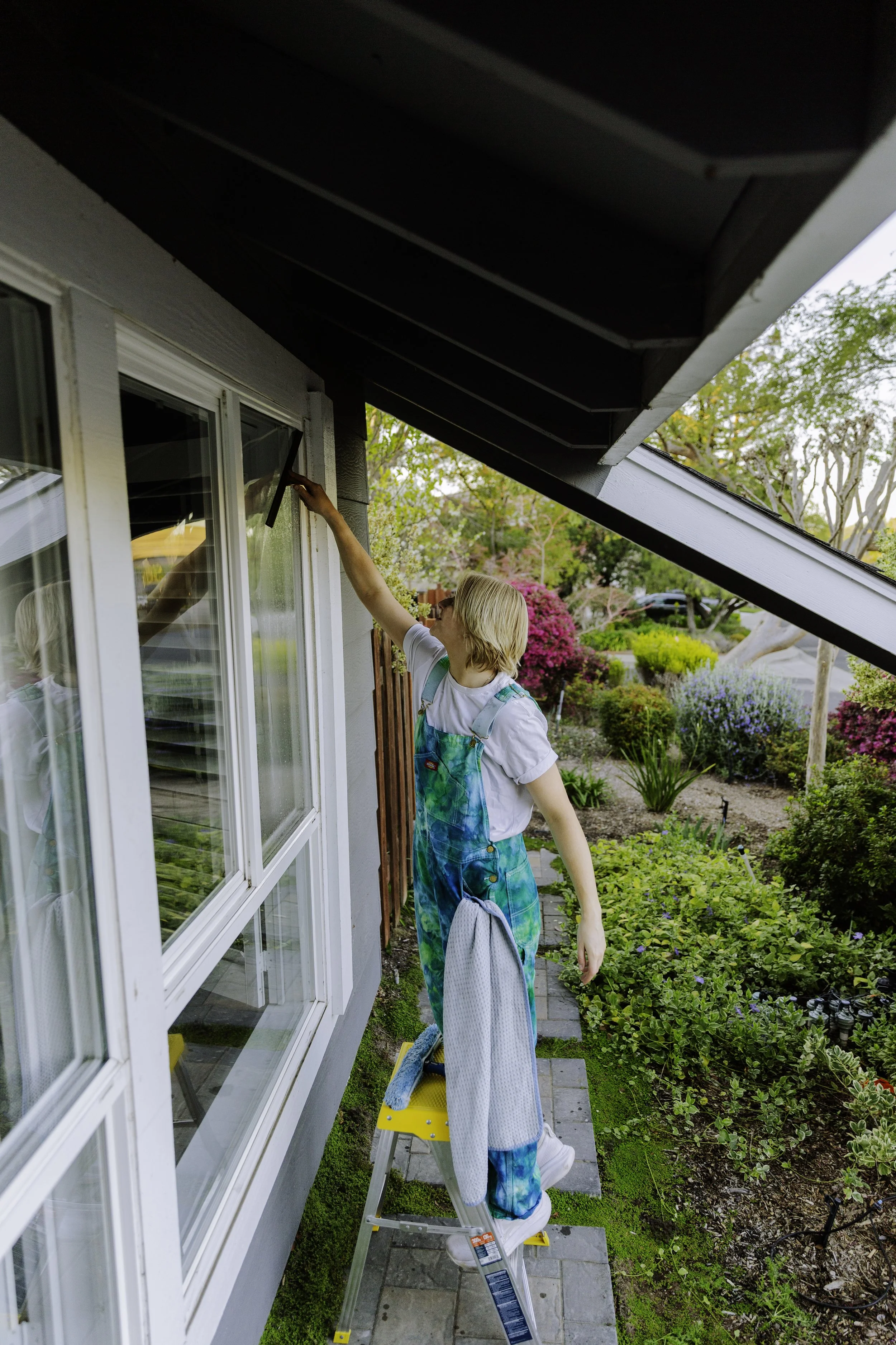 A woman standing on a yellow step stool cleaning the exterior window of a house in a lush garden with colorful bushes and trees.