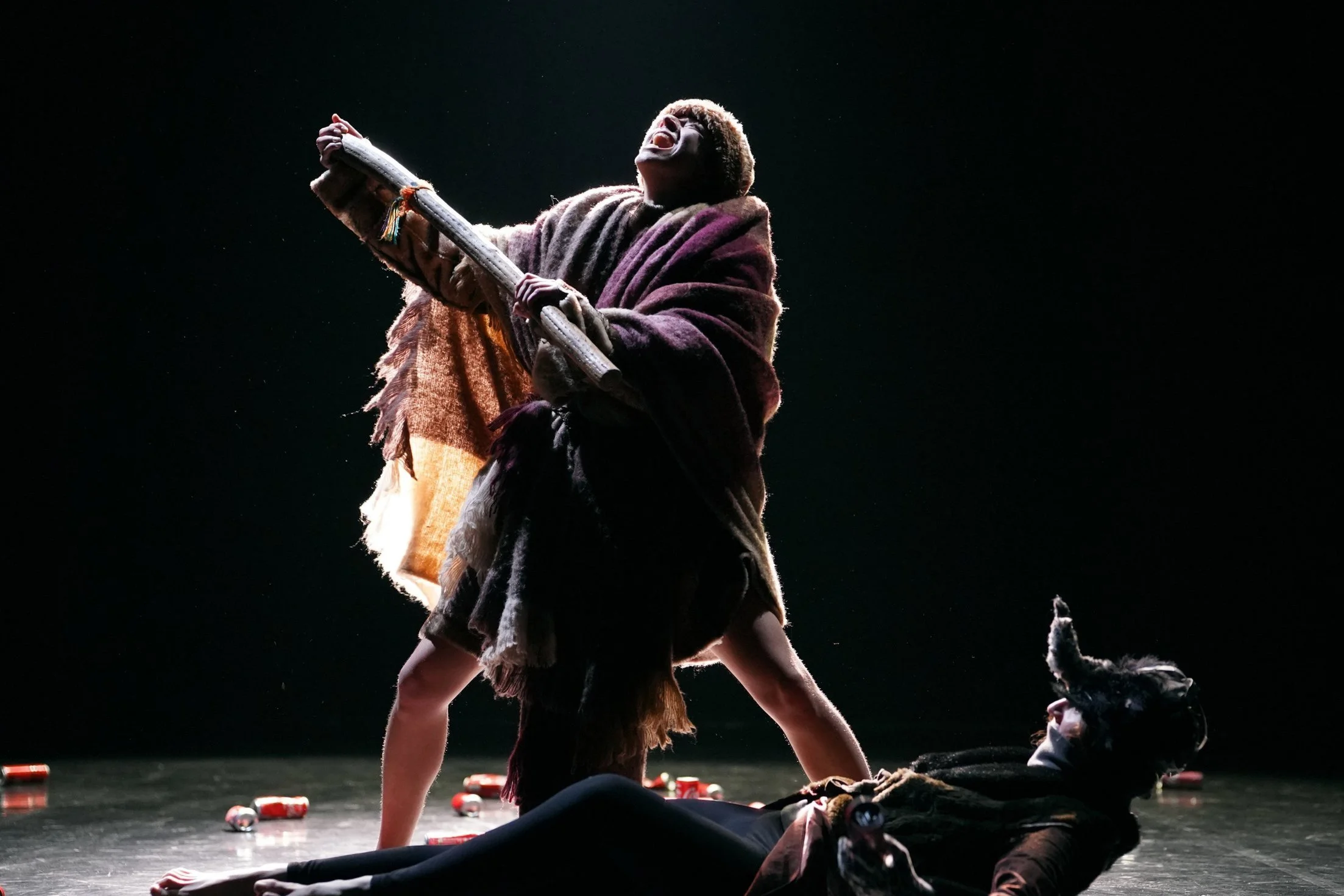 Une scène de théâtre avec deux acteurs en costumes, dont un portant un masque de chien, engagés dans une performance expressive, sur une scène sombre avec des canettes au sol.