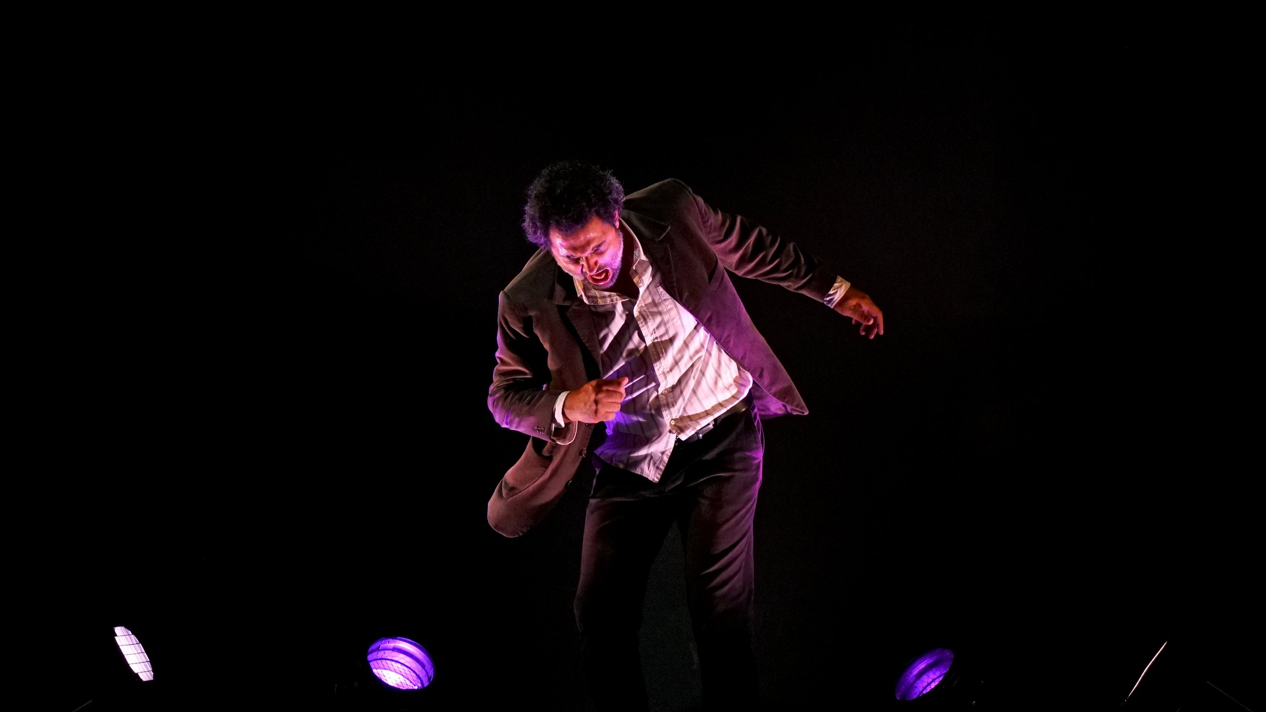 Un homme en costume marron et chemise blanche, chantant ou exprimant une émotion forte sur scène, éclairé par des lumières violettes dans un environnement sombre. Extrait de la pièce Où est mon chandail islandais?