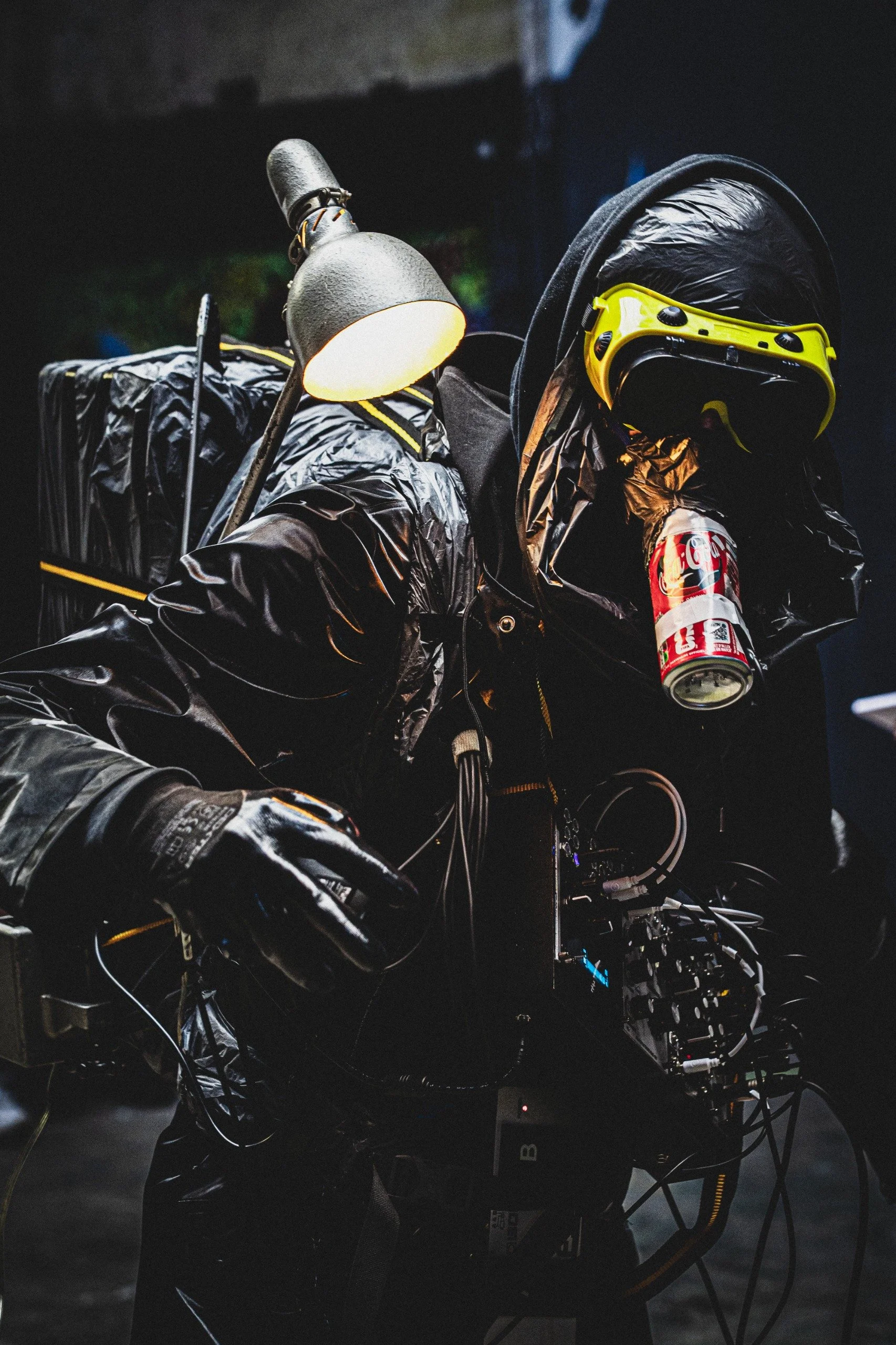 Une scène sombre avec des équipements électroniques, un casque de réalité virtuelle jaune, une lampe de bureau, des câbles et un canette de Coca-Cola, suggérant un environnement de création ou de développement technologique. Extrait de Ultra-Life.