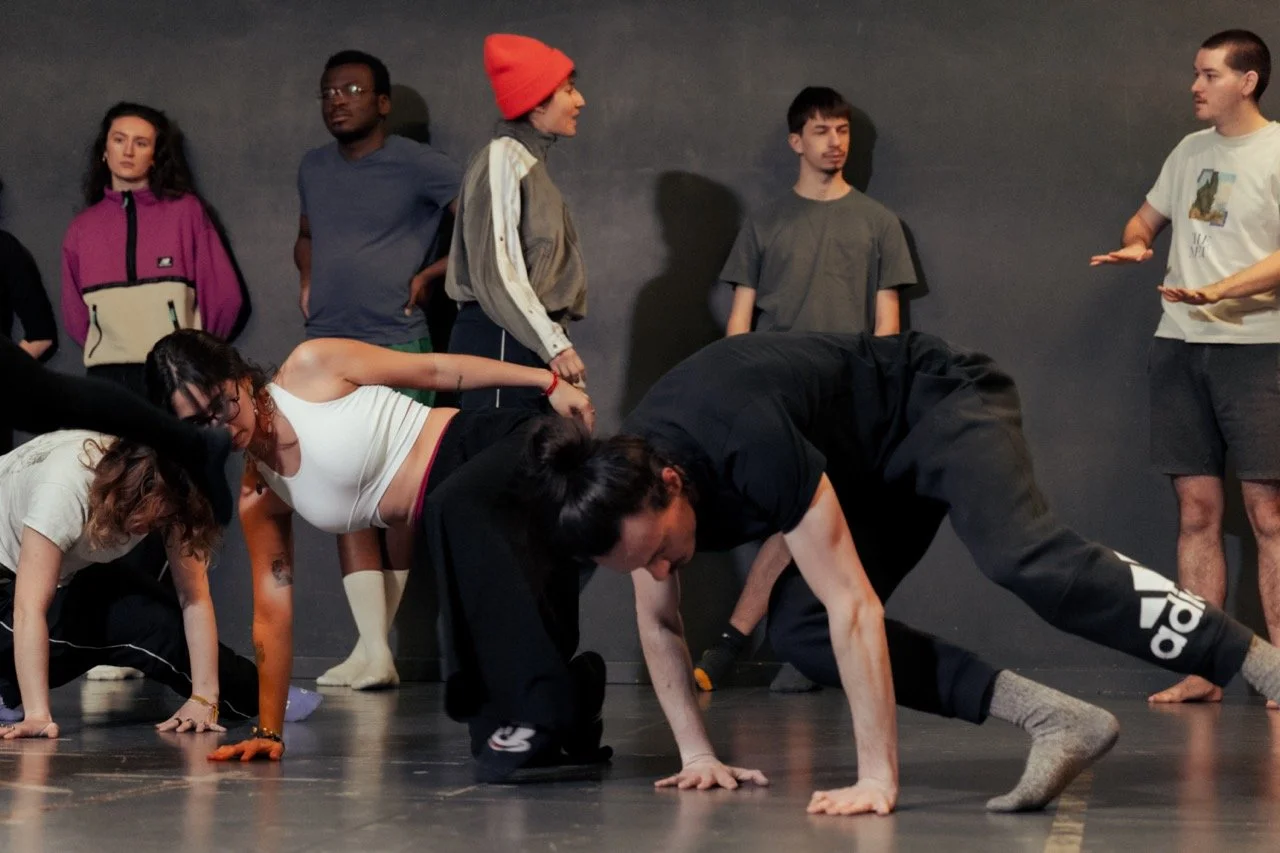 Groupe de personnes participant à une séance de danse ou de théâtre, avec deux personnes à l'avant en position de pognée au sol et d'autres debout en arrière-plan, dans une pièce sombre.