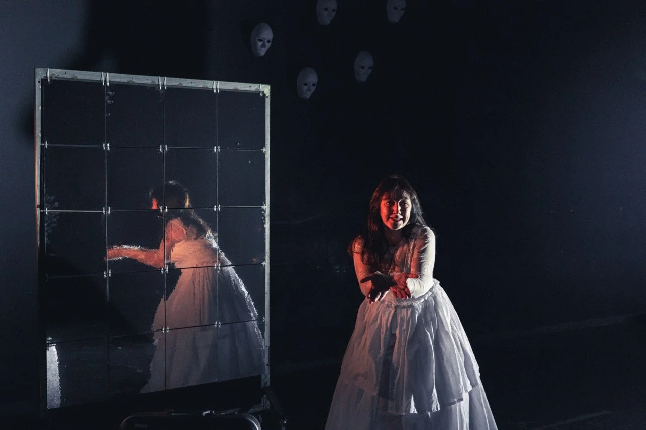 Une jeune fille en robe blanche se tient devant un miroir, avec un fond sombre et des masques blancs accroché au mur au-dessus dans le cadre de la formation mise en scène