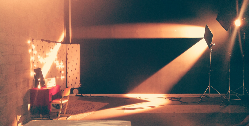 Un mur d'enceinte avec un spot lumineux en studio photo, un fond noir, deux lampes sur trépied et une table avec une décoration lumineuse en arrière-plan.
