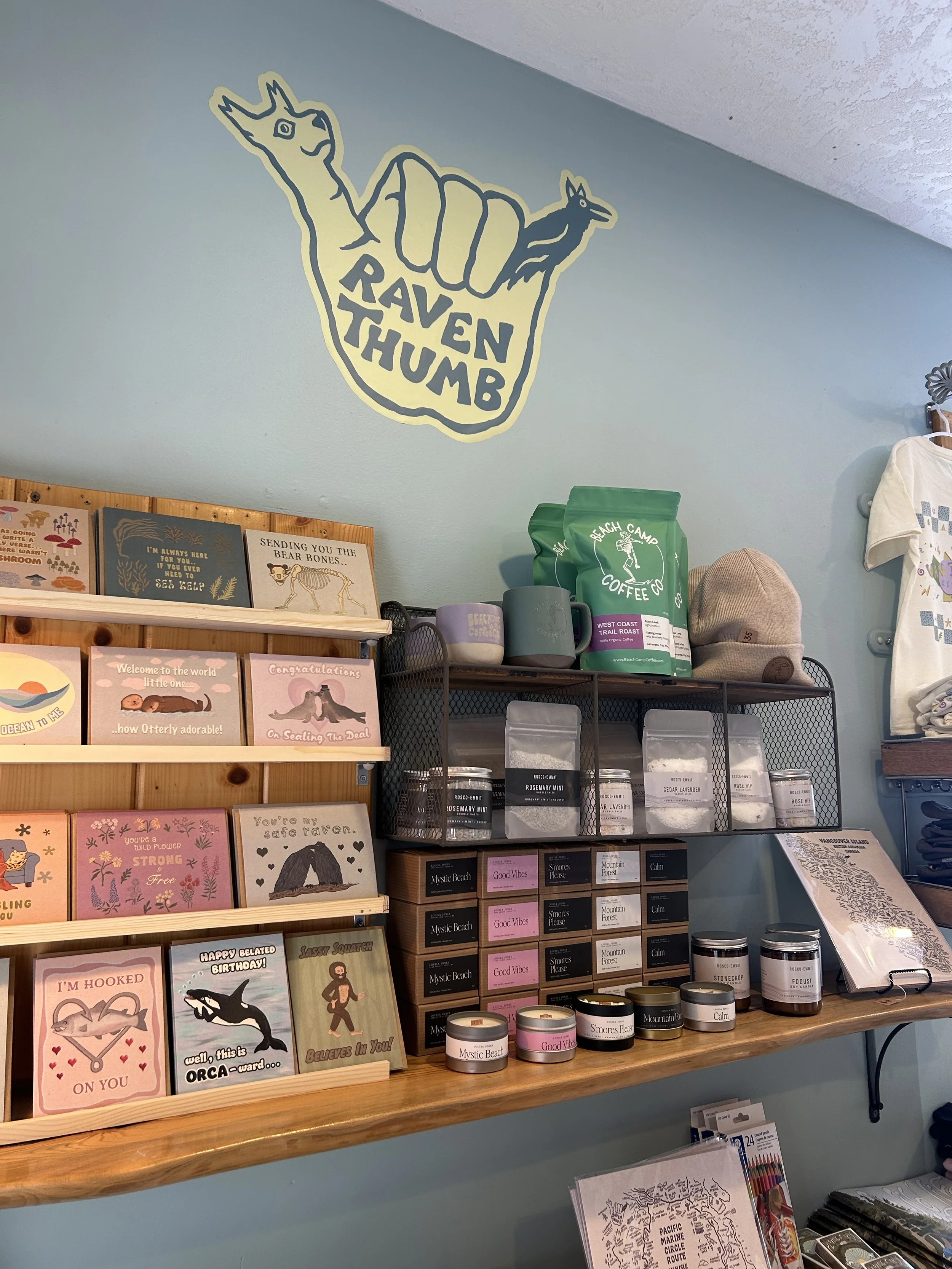 A wall with a mural of a fist holding a llama and a fox, with the words "RAVEN THUMB" inside the fist. Below, shelves display greeting cards, tea, and other products in a store.