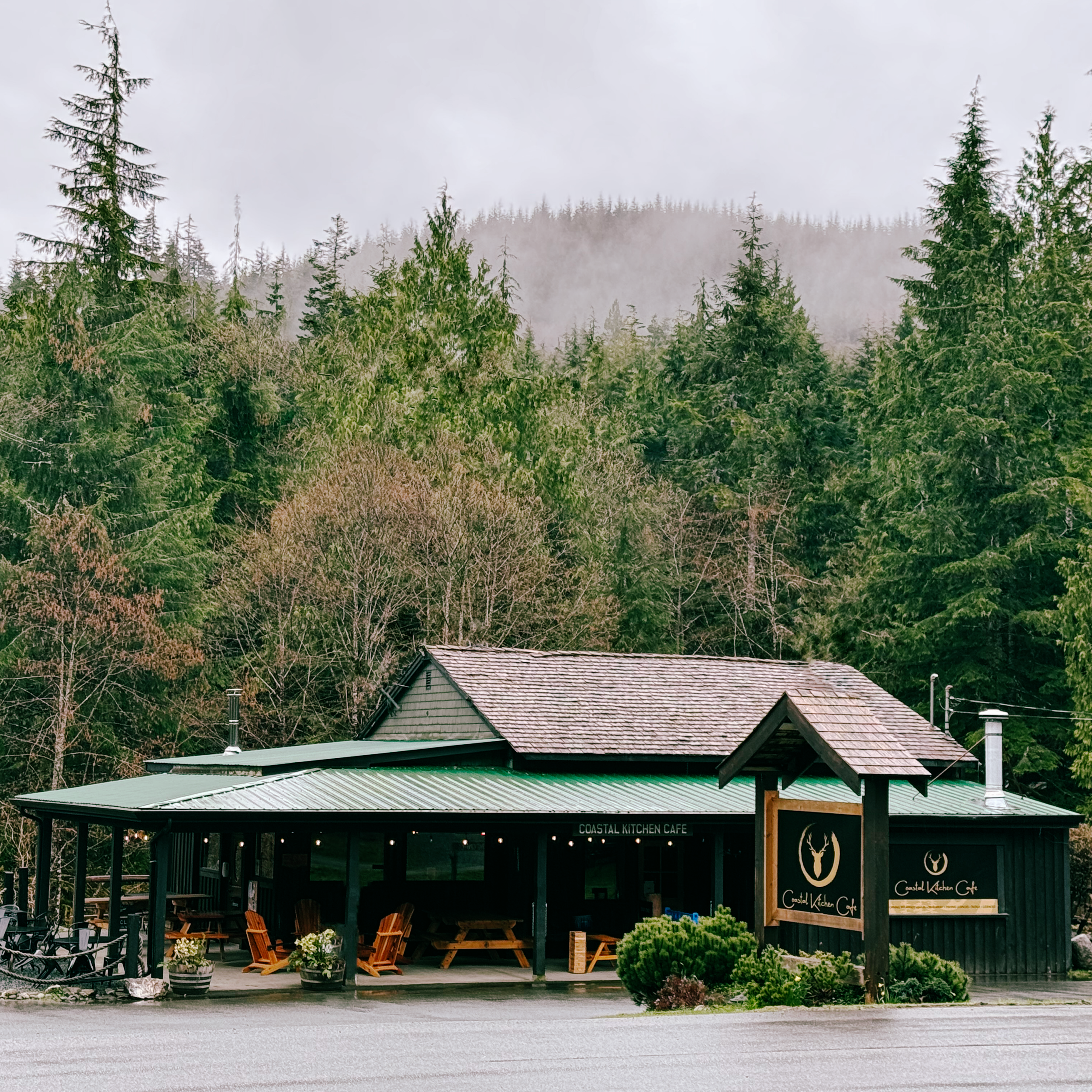 A rustic cafe with outdoor seating, surrounded by lush green trees and mountains in the background.
