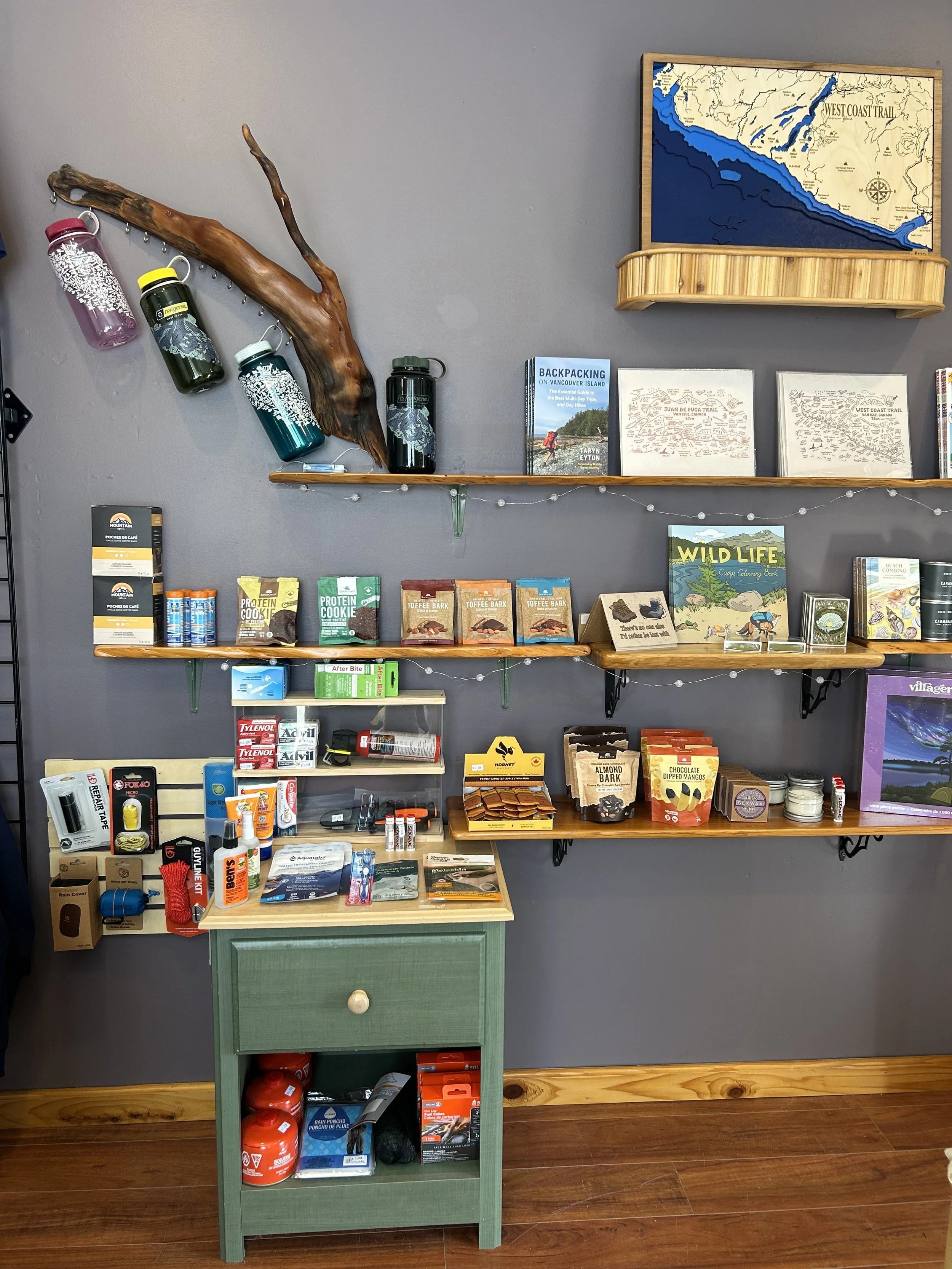 A store display with various outdoor and hiking gear, including water bottles, maps, books, snacks, first aid supplies, and small tools, arranged on shelves and a green stand against a gray wall.