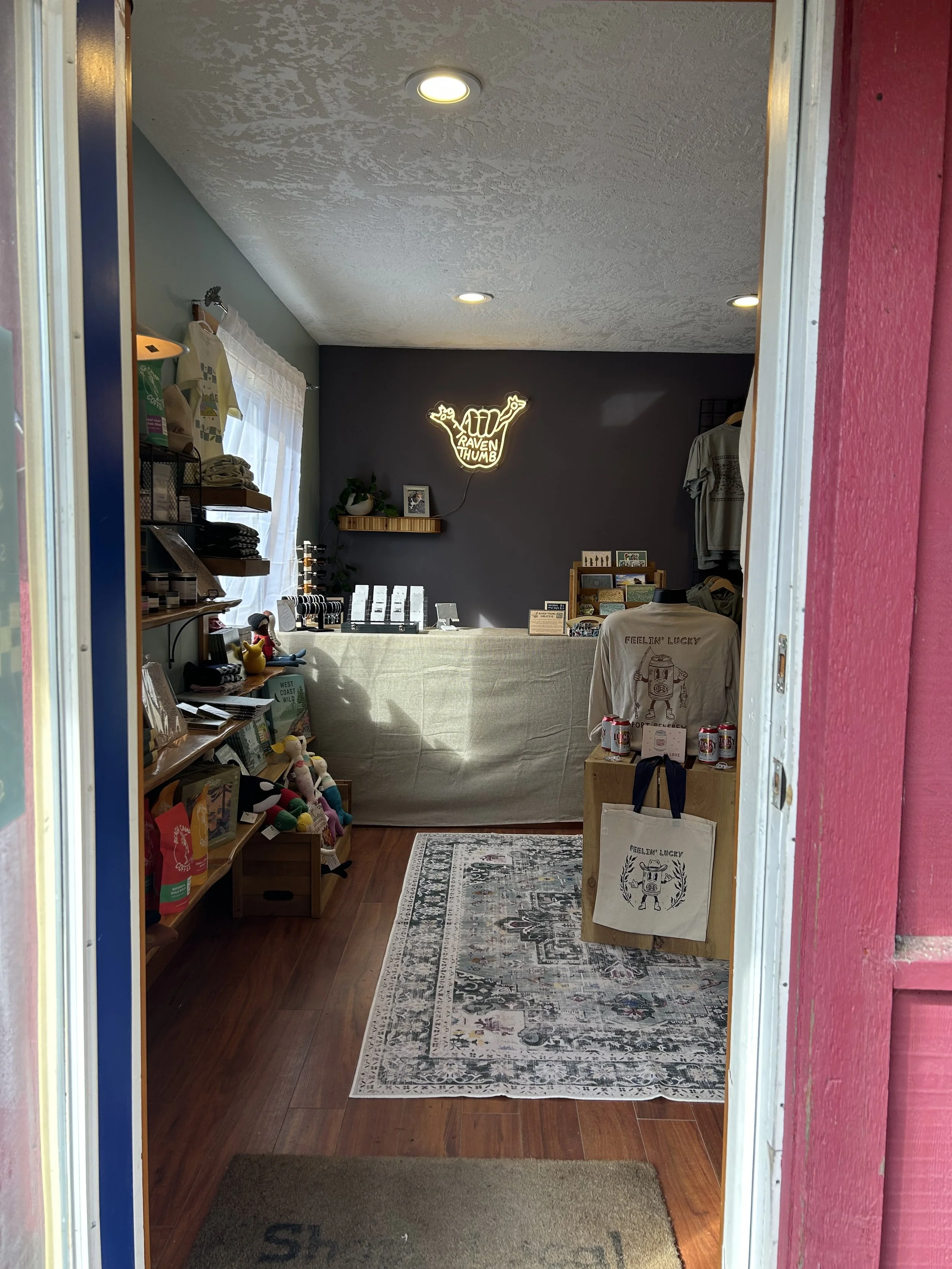 Interior view of a shop selling various items, featuring a dark gray wall with a neon sign that says 'AT&T Raven Thumb', shelves with books and collectibles on the left, a table with items in the center, a small plant on a wall shelf, and a rolling rack with clothes on the right.