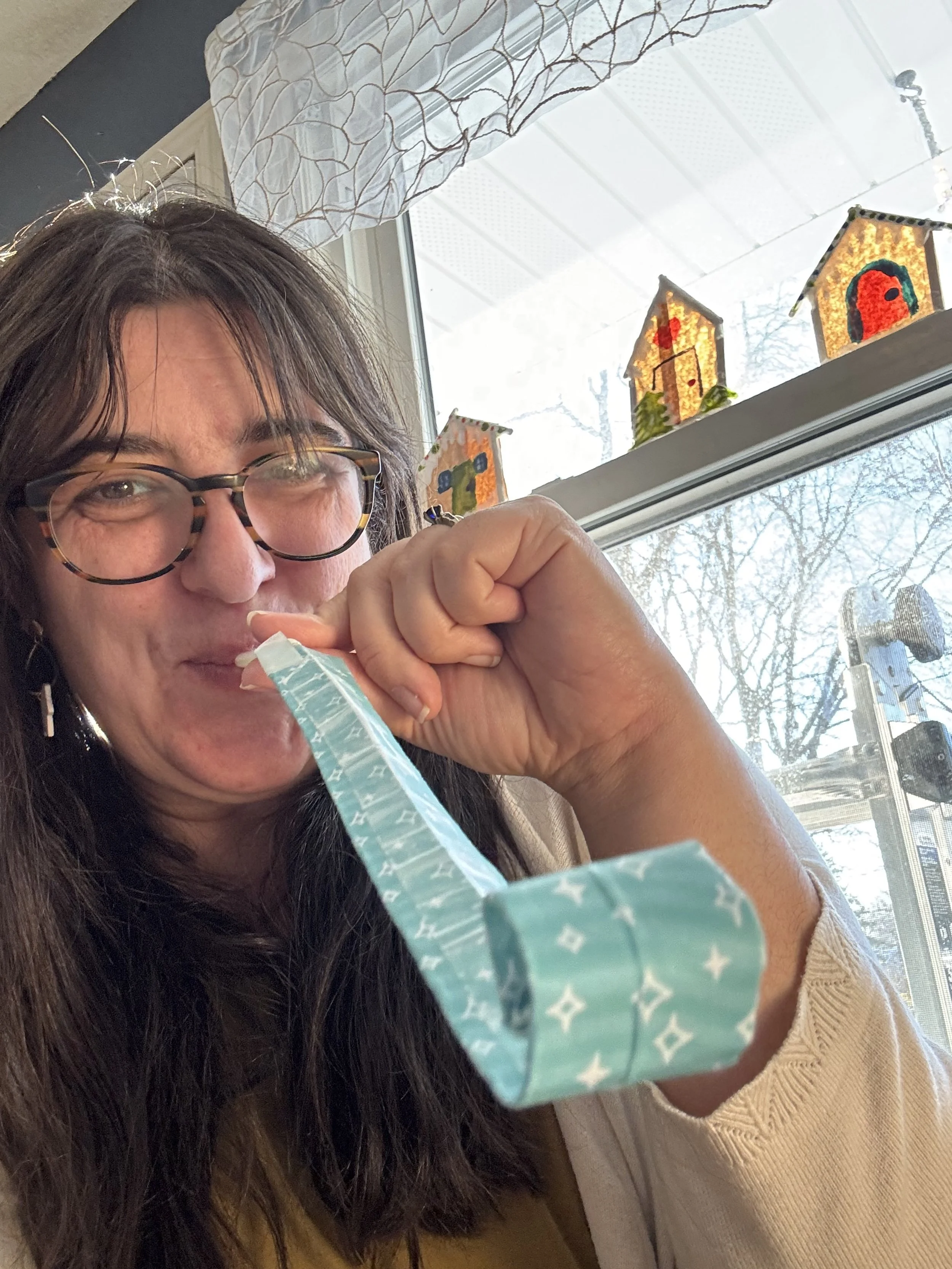 A woman blowing a paper party blower with popsicle stick gingerbread house crafts in the background.