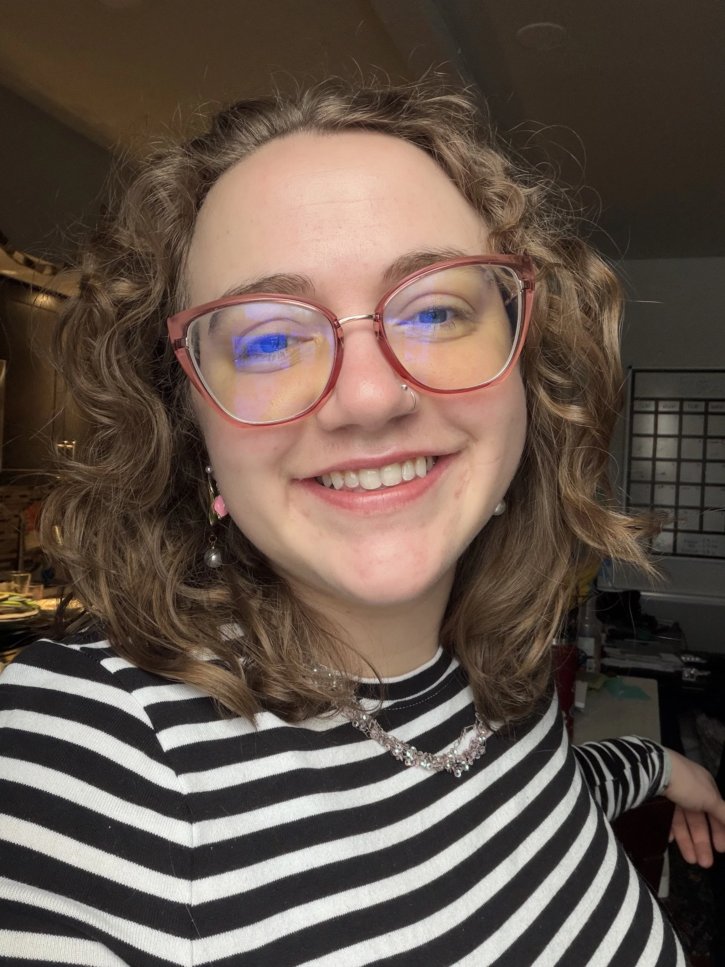 Piper Burris smiling - curly, shoulder-length blonde hair, pink glasses, black and white striped shirt
