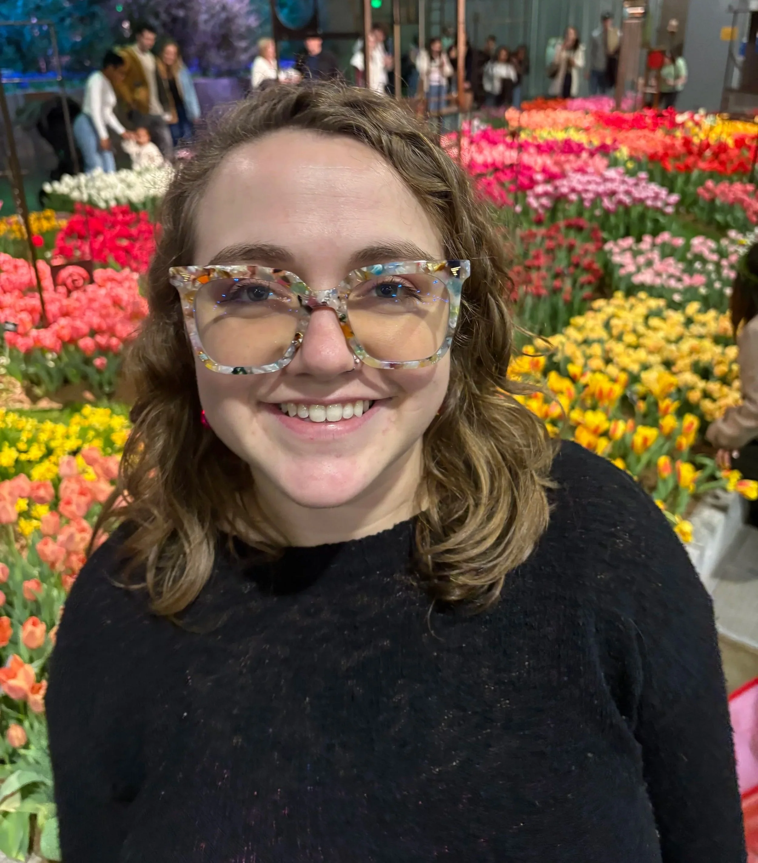 Photo of Piper Burris - blonde, shoulder-length hair, black sweater, colorful glasses. She is in focus and there are many different colored tulips behind her - pink, yellow, red, peach, white, to name a few.