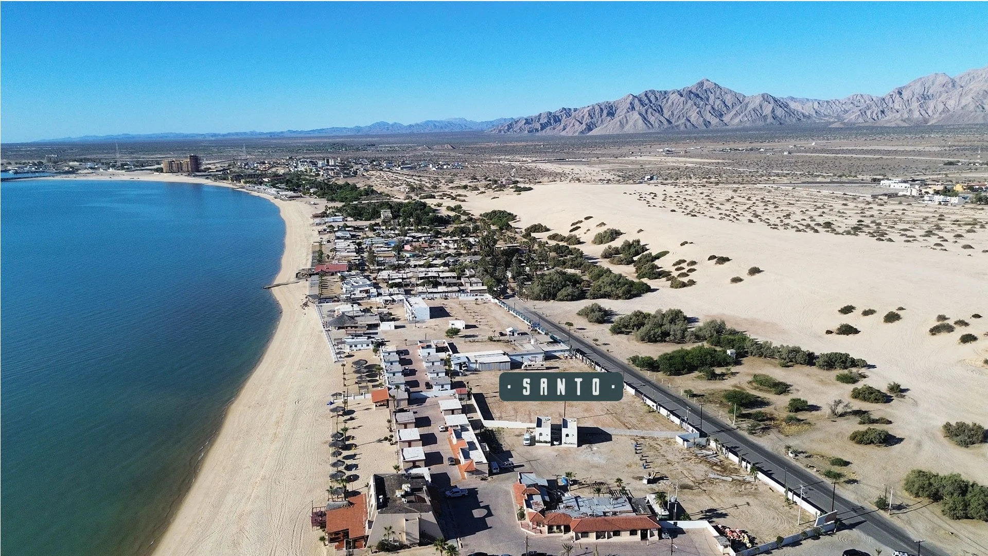 Aerial view of a coastal town with a sandy beach and calm water, surrounded by desert landscape with mountains in the background, located in Santo.