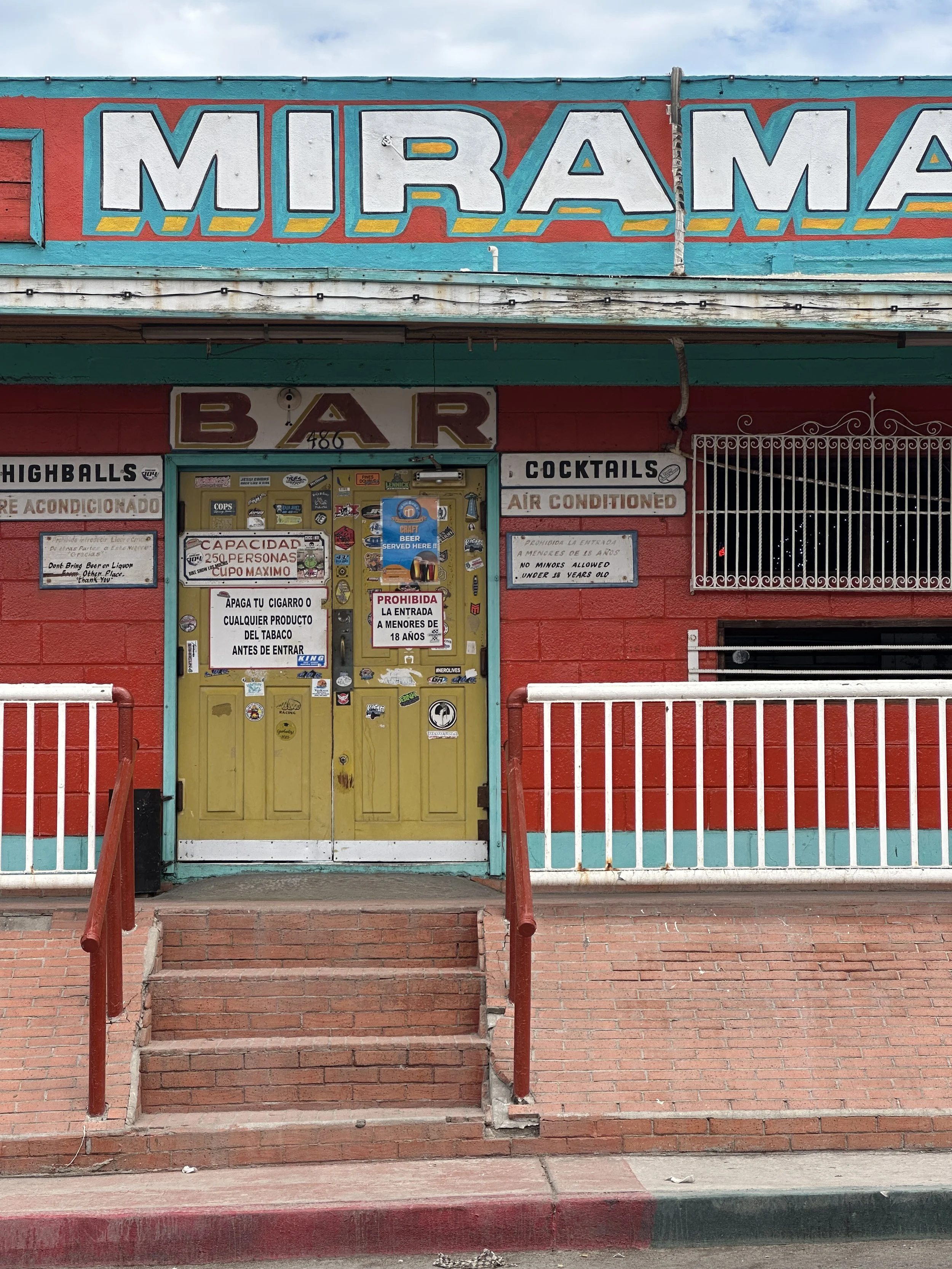 Front entrance of a building with colorful signs and stickers. The main sign displays in large white letters with teal outlines that say "MIRAM" on a red background. Below, a smaller sign indicates the entrance is for a bar and lists amenities such as highballs, cocktails, and air conditioning. The door and window are covered with various stickers and signs, some warning about no minors allowed and no bringing alcohol or tobacco before entry. Red and white railings are positioned in front of the stairs leading to the door, and brick flooring extends to the sidewalk.