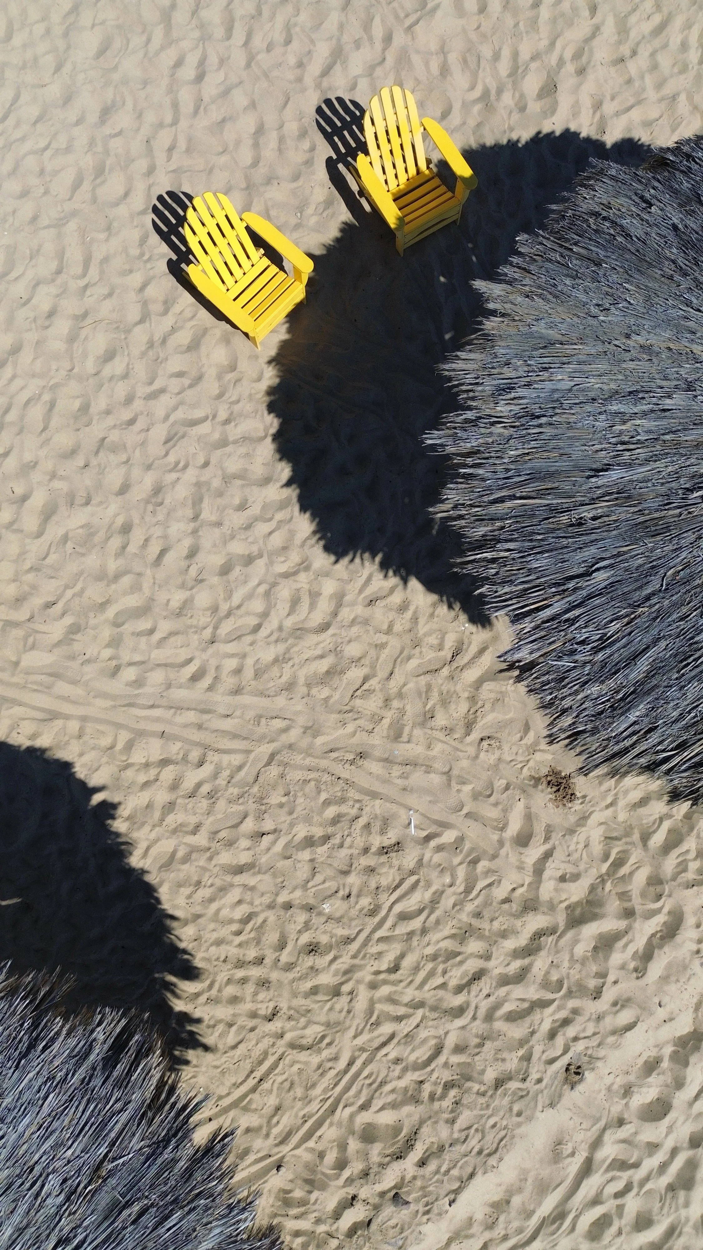 Two yellow Adirondack chairs on sandy beach seen from above, with shadows and a thatched roof in the foreground in SANTO san felipe