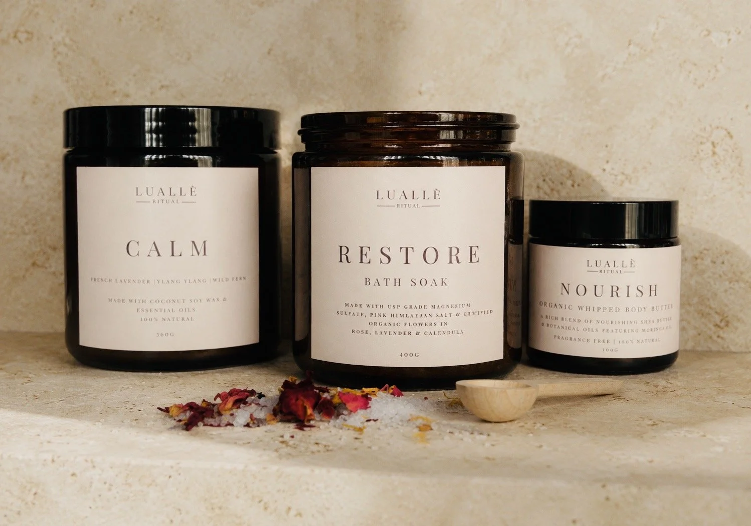 Three amber jars labeled 'Calm,' 'Restore,' and 'Nourish' arranged on a beige surface with a small wooden scoop and flower petals scattered in front. Background is a textured beige wall.