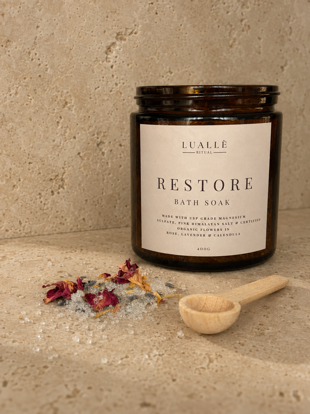 A jar of Luallë Ritual Restore bath soak on a beige countertop. Next to it, there is a small wooden scoop and a pile of bath salts mixed with dried flower petals.
