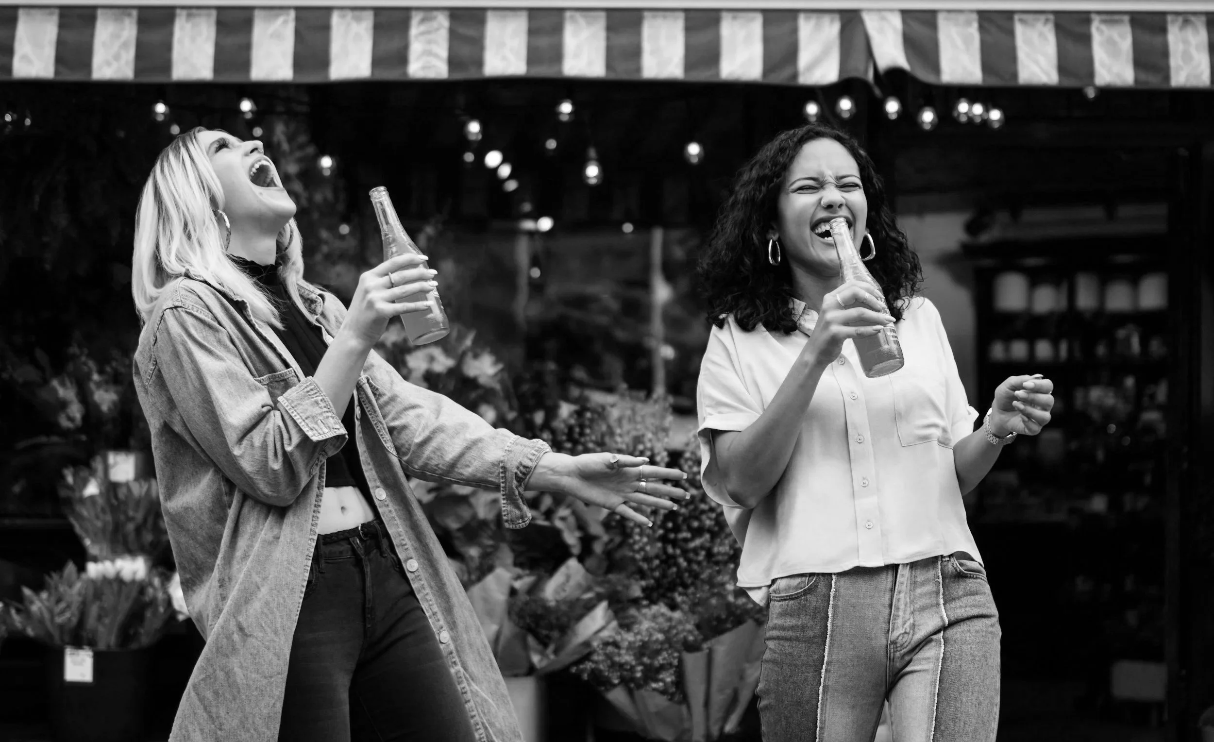 Zwei lachende Frauen, eine mit blonden Haaren und die andere mit dunklen, trinken Bier in einem Blumenladen oder Markt im schwarzen und weißen Foto.