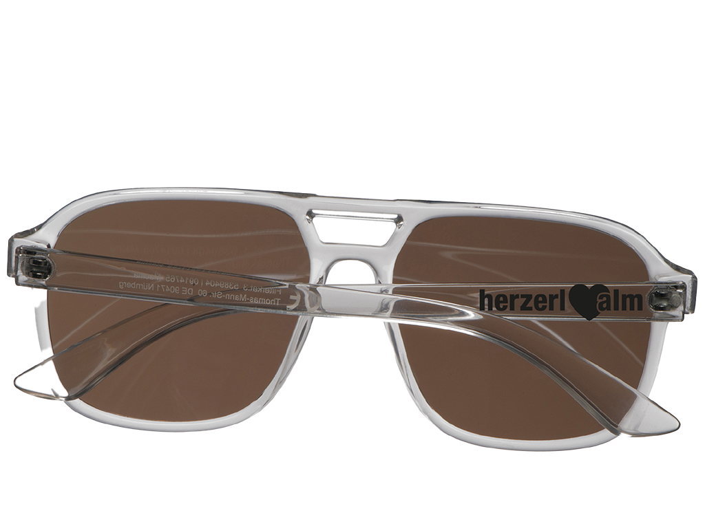 Sonnenbrille mit braunen Gläsern und transparentem Rahmen, mit Herzsymbol und Text "Herzer|alm" auf der Seite.