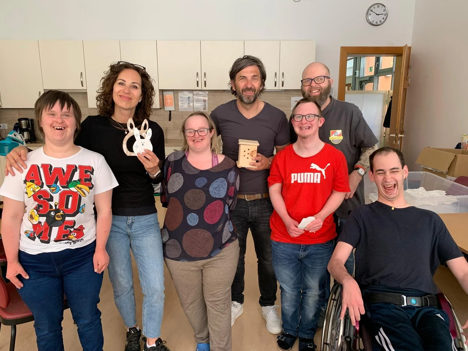 Eine Gruppe von acht Personen, einige mit körperlichen Beeinträchtigungen, stehen in einer Küche und lachen. Zwei Frauen und sechs Männer, einige halten Geschenke.