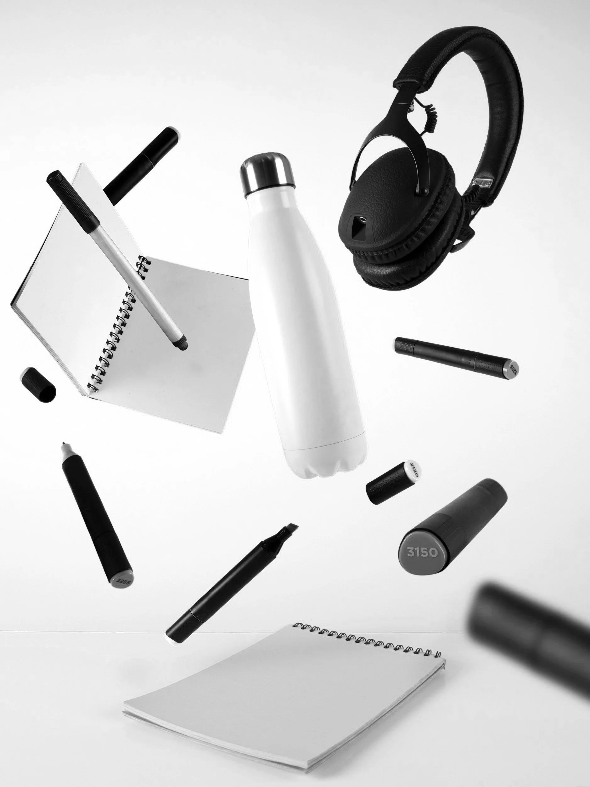 Schwarz-weiße Aufnahme von verschiedenen Büromaterialien und Gegenständen, darunter ein Notizbuch, Wasserflasche, Kopfhörer und Marker, die in die Luft zu schweben scheinen.