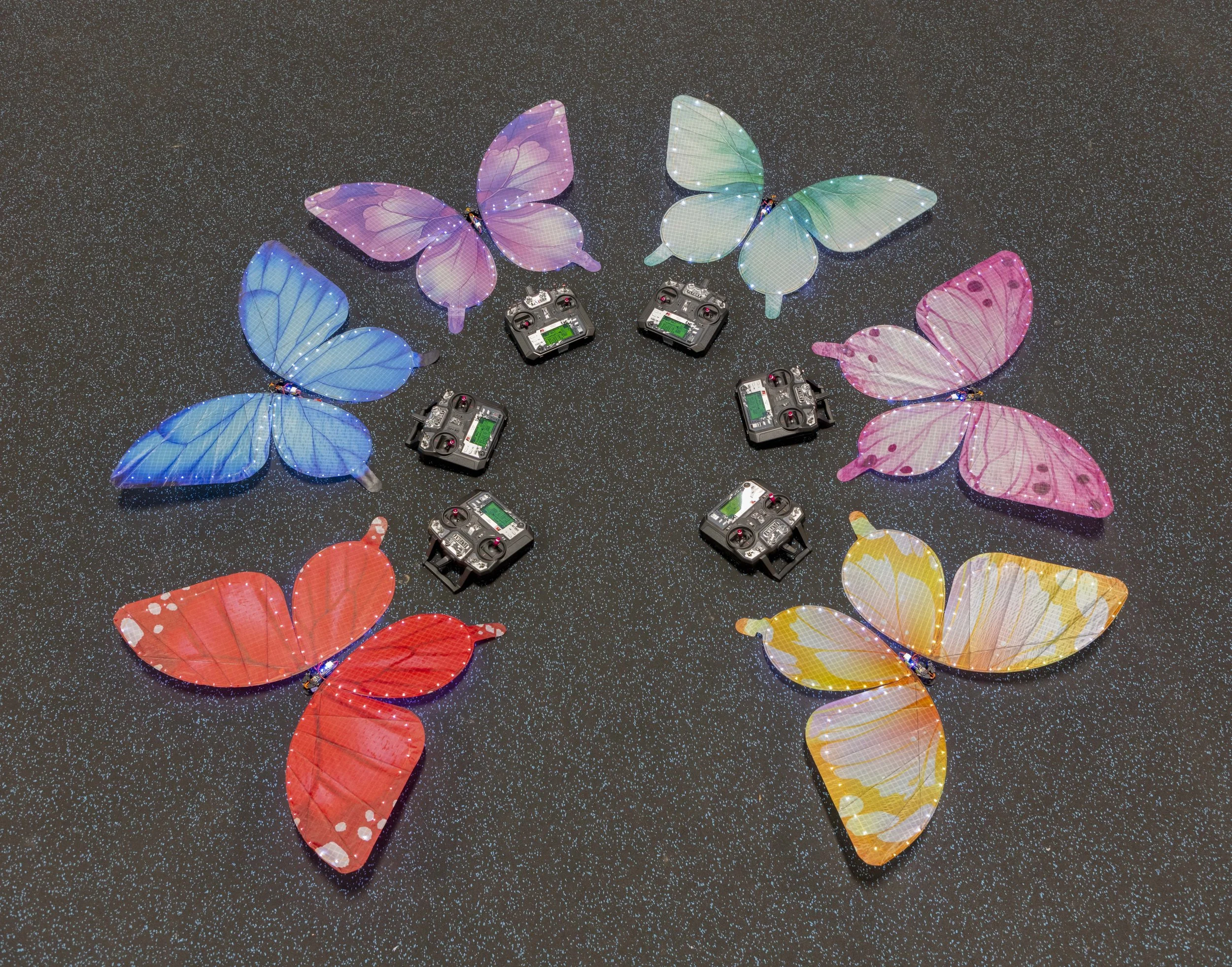 Eight colorful butterfly-shaped LED wings arranged in a circle on a glittery black surface, with five remote controls placed among them.