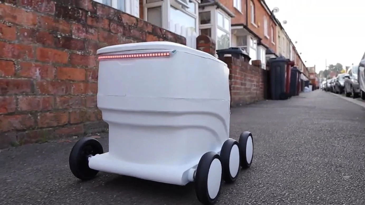 A white delivery robot with four black wheels on a sidewalk in front of a brick wall and residential buildings.