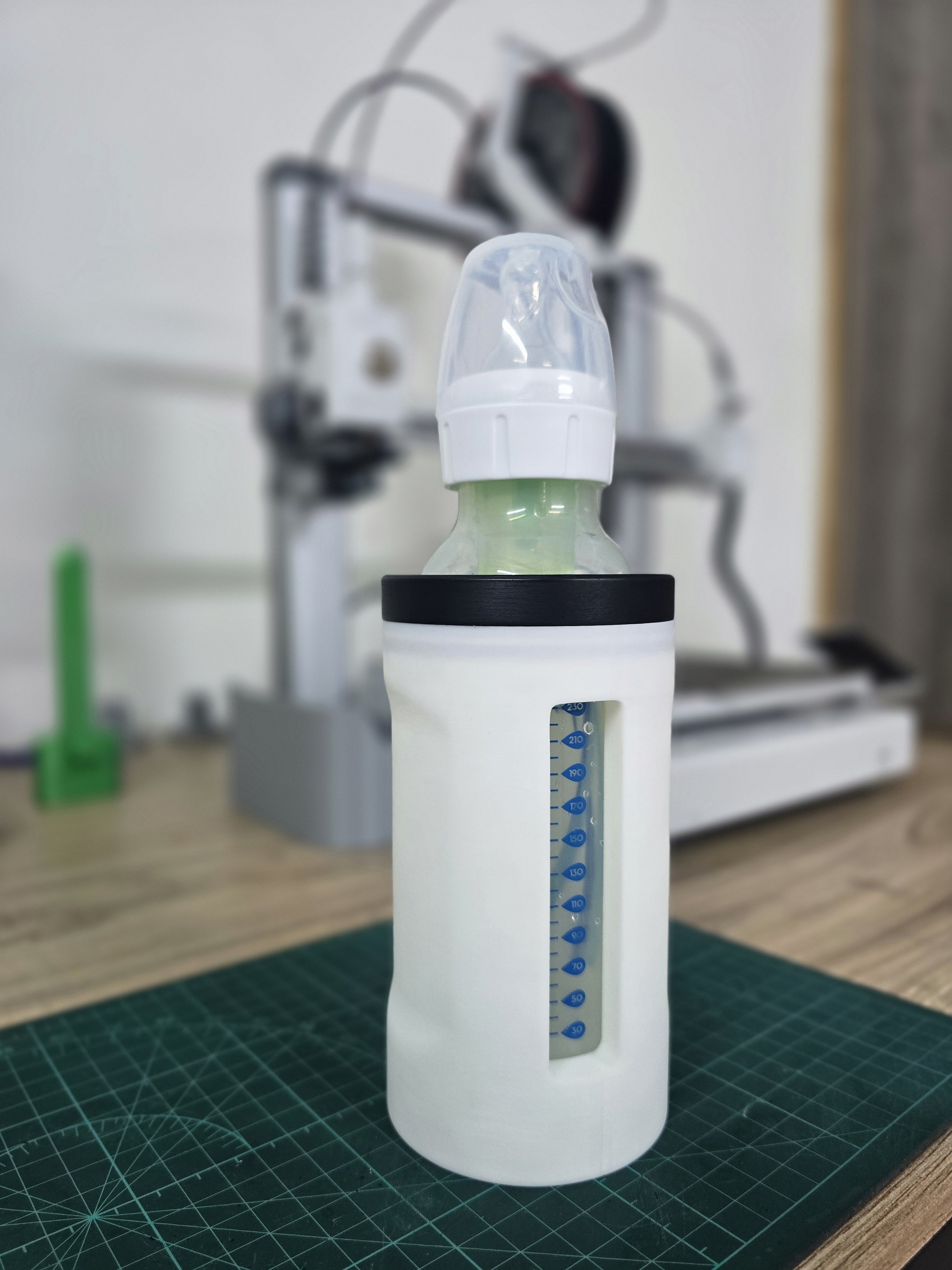 A baby bottle with a clear plastic nipple, green liquid, and a white sleeve with a water level indicator, placed on a green cutting mat on a wooden table.