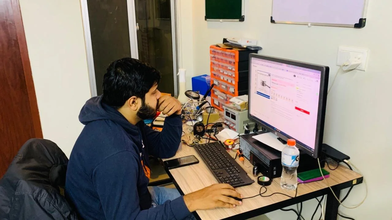 Young man working at a cluttered desk with a computer, monitor displaying a webpage, and various electronic components and tools.