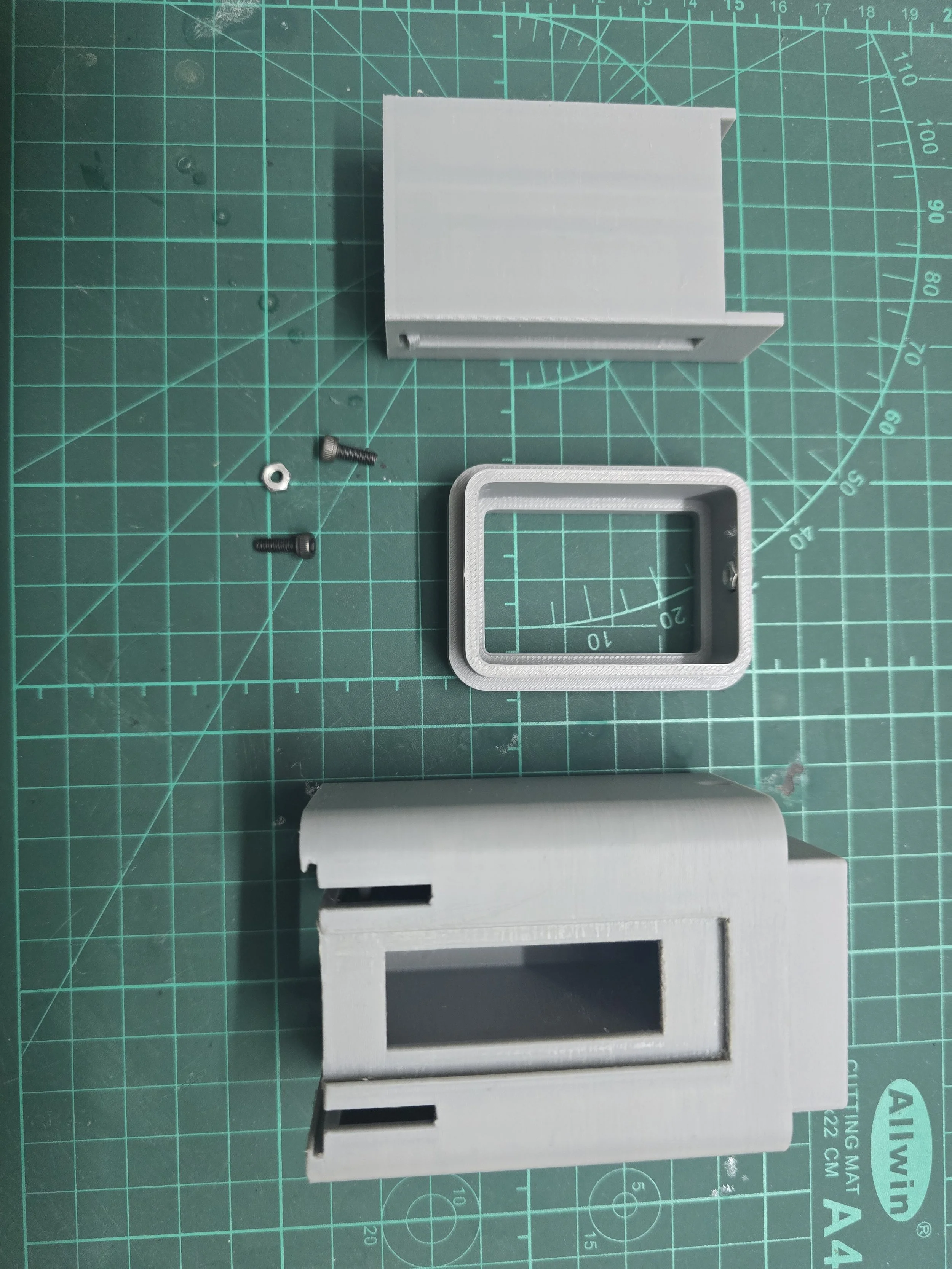 Disassembled parts of a small electronic project or enclosure, including a white plastic box, a metallic frame, a white rectangular cover, screws, and a nut, placed on a green cutting mat with measurement markings.