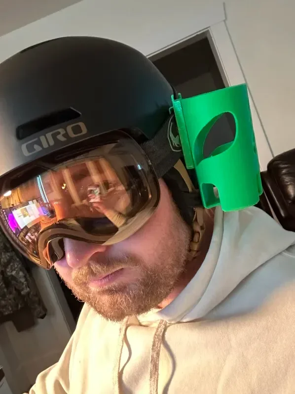A man wearing a black Giro helmet with a green face shield attached, and large ski or snowboarding goggles. He is indoors and has a beige hoodie on.