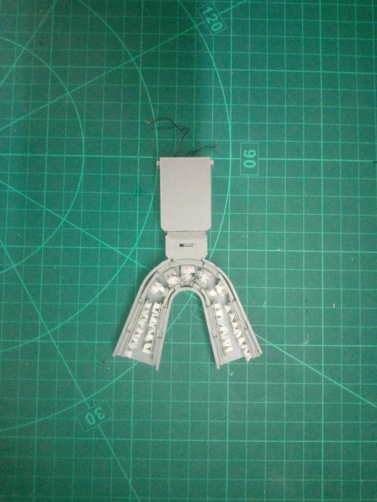 A small white plastic model of a horseshoe magnet with an attached rectangular component and wires, placed on a green cutting mat with grid lines and measurement markings.
