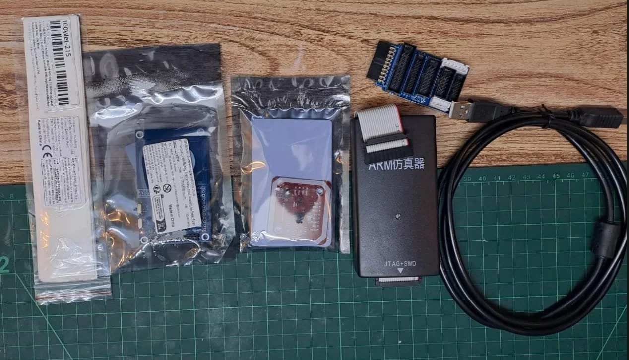 Electronic components and tools on a wooden surface, including a green cutting mat, a cooling fan, a hard drive, an ARM emulator, a USB cable, and connectors.
