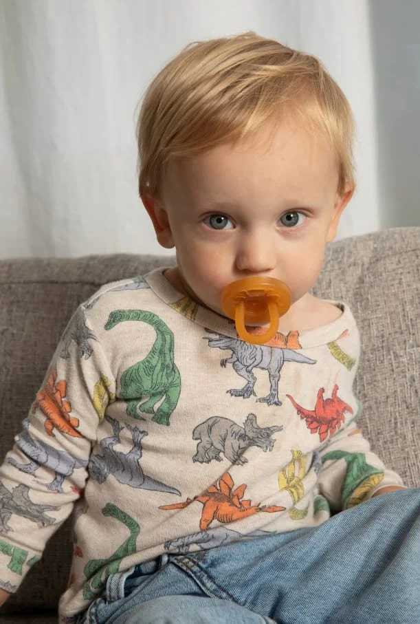 A young child with blond hair and blue eyes sitting on a couch, wearing a dinosaur-printed shirt and an orange pacifier in his mouth.