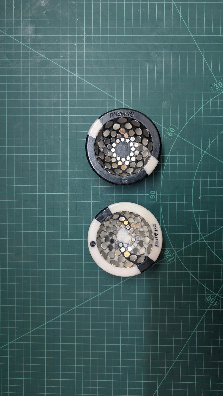 Two metal herb grinder parts, one black and one white, with perforated surfaces, on a green cutting mat with measurement markings.