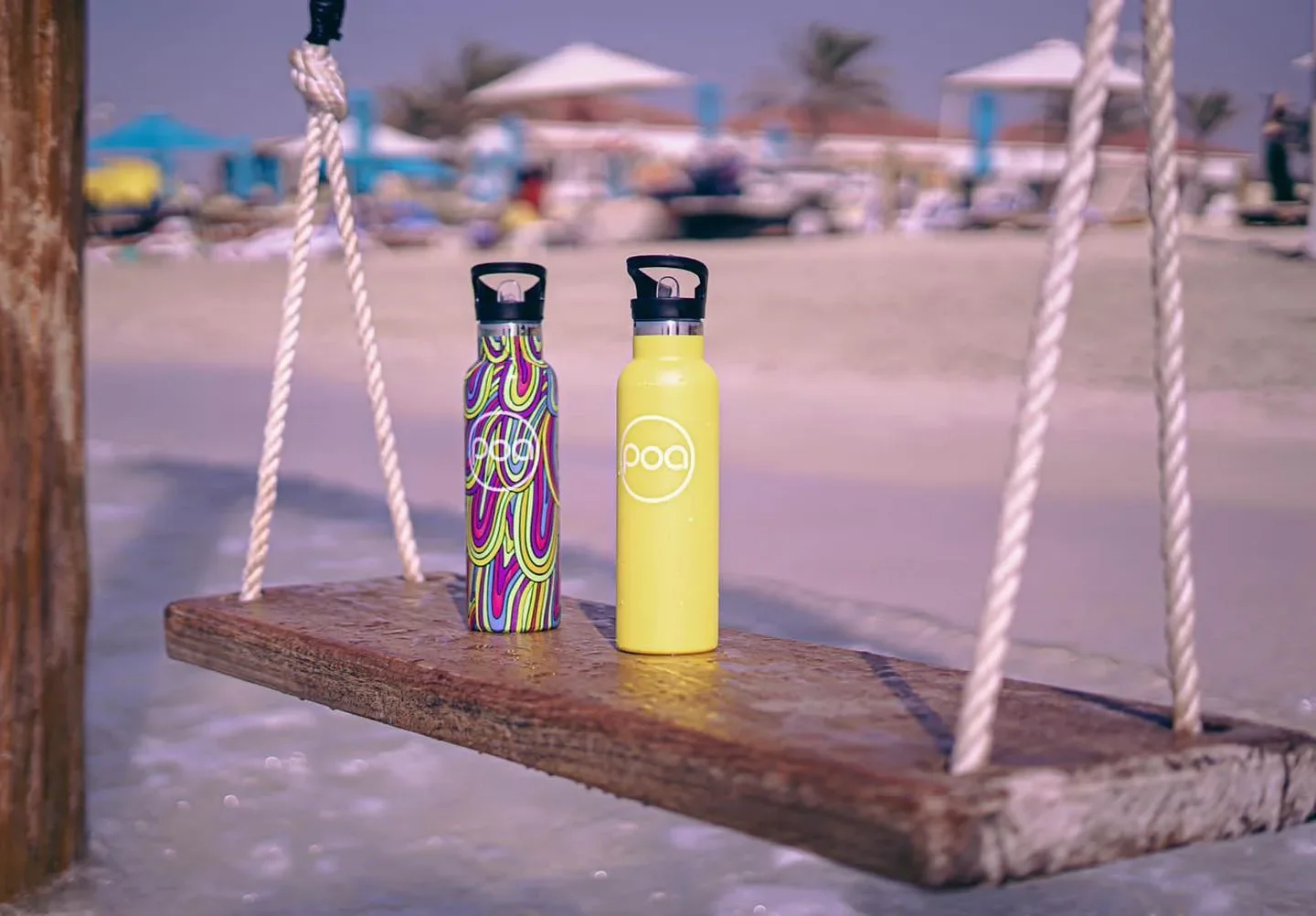 Two water bottles, one with a psychedelic swirl pattern and the other yellow, placed on a wooden swing at the beach with umbrellas and beachgoers in the background.