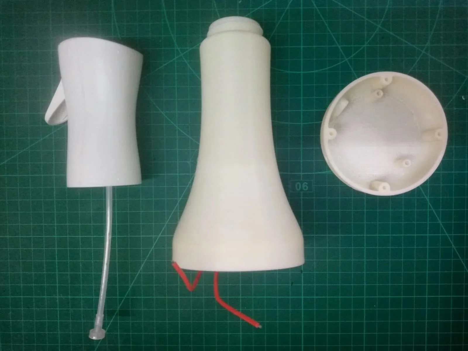 Disassembled plastic lamp parts including a white lampshade with a transparent support and a red wire, a large tapered white base with red wires, and a circular white top cover with screw holes, all placed on a green cutting mat.