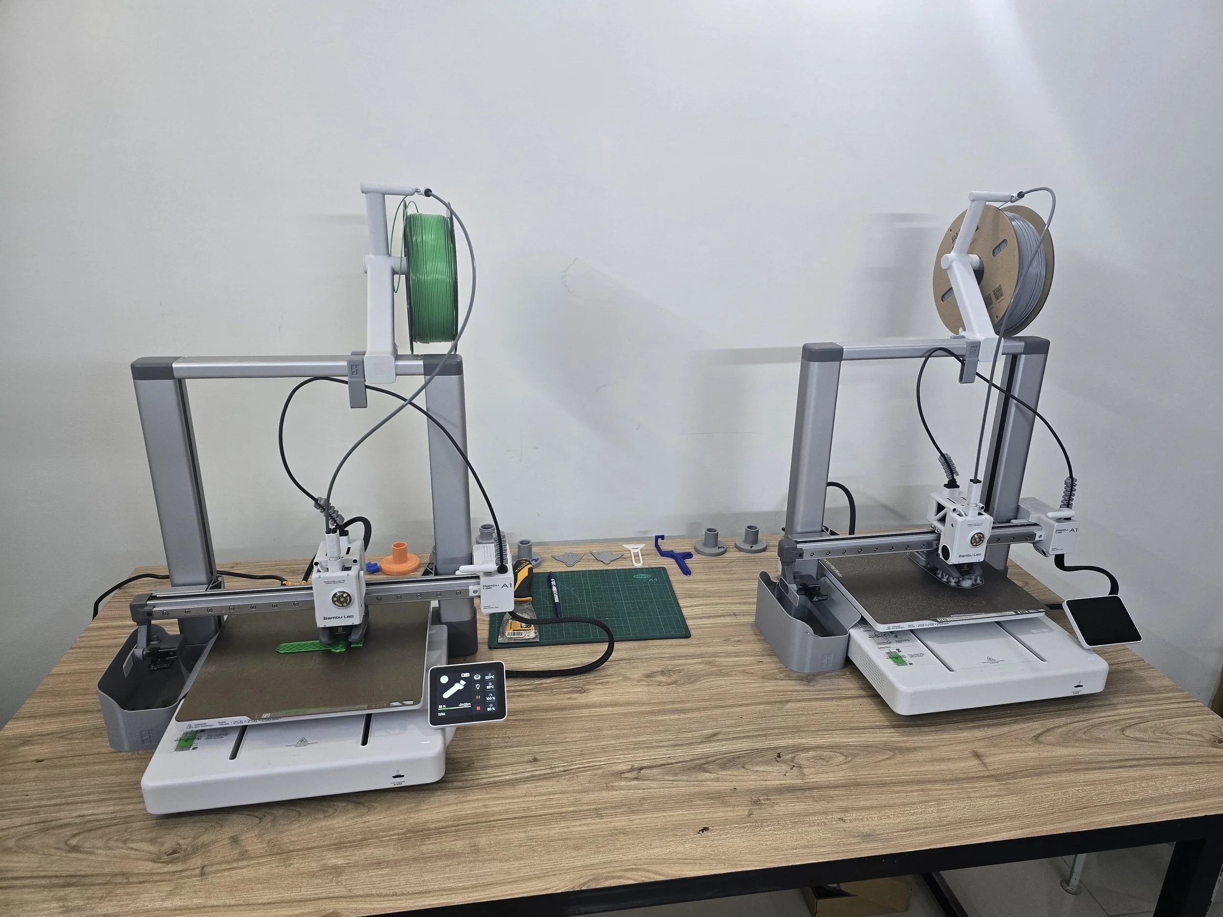 Two 3D printers on a wooden table, each with a spool of filament; one with green filament and the other with gray filament. Tools and a cutting mat are on the table.