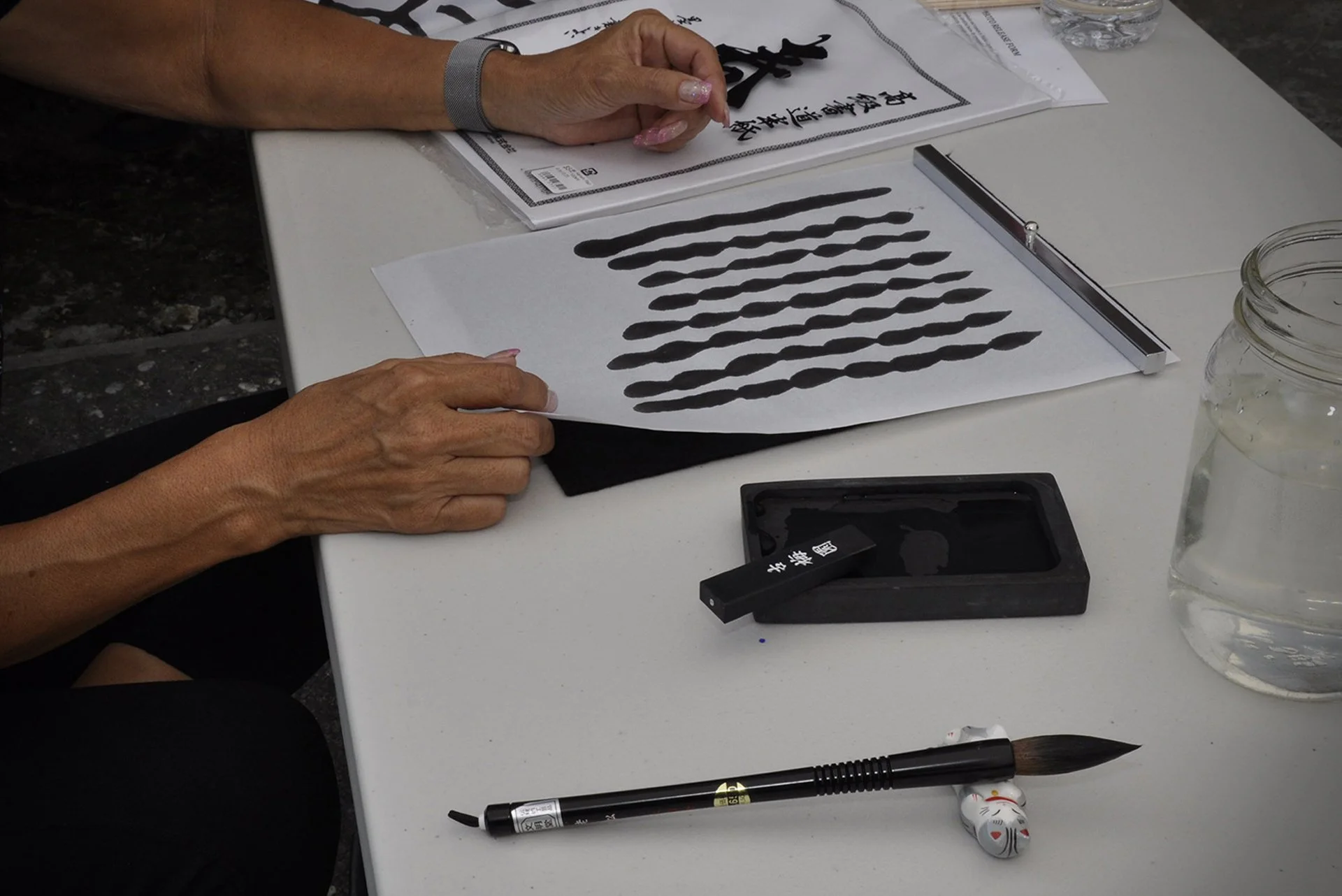 Person demonstrating traditional Asian ink brush strokes on paper, with tools including ink, brush, and paper on a white table.