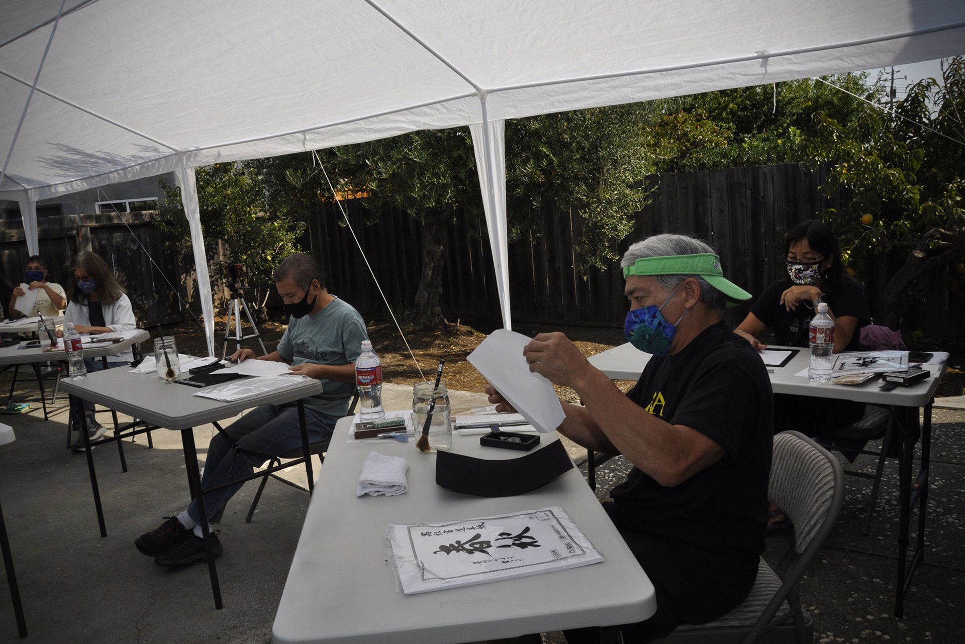 People sitting at tables under a large white canopy outdoors, reading and examining papers, wearing face masks.