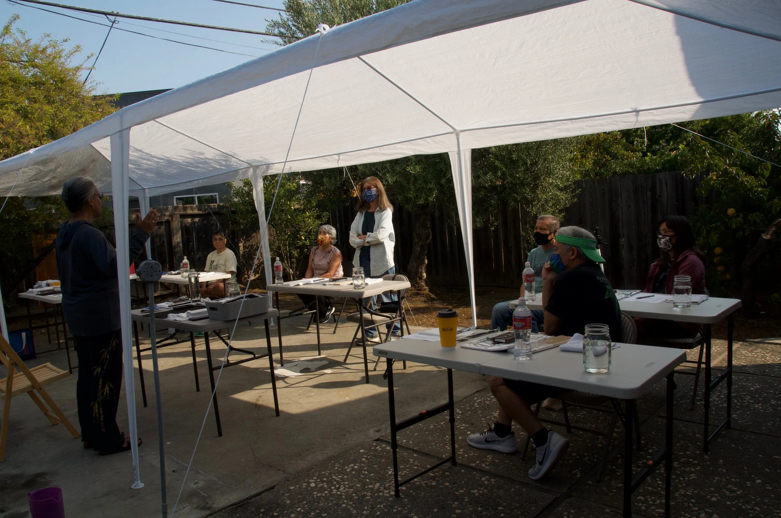 A group of people gathered under a white canopy tent outdoors, participating in a meeting or workshop with a woman speaking at the front. They are seated at tables with water bottles, notebooks, and drinks. All individuals are wearing face masks.
